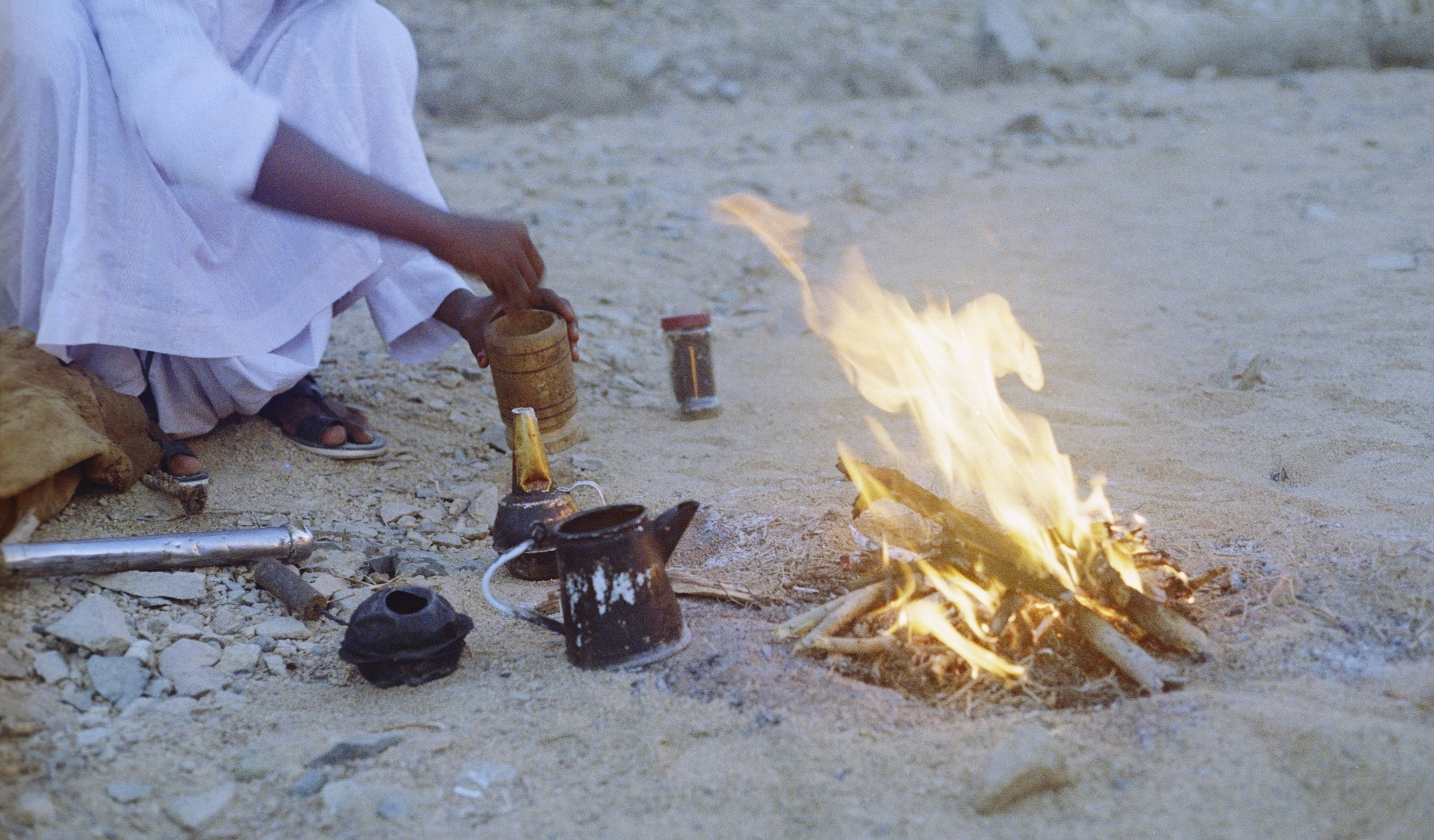 A person in a white robe preparing coffee by a small campfire on sand