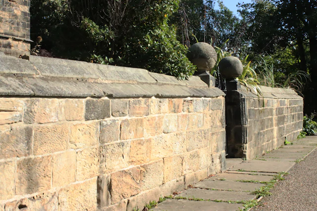 A weathered stone wall with spherical finials beside a paved sidewalk
