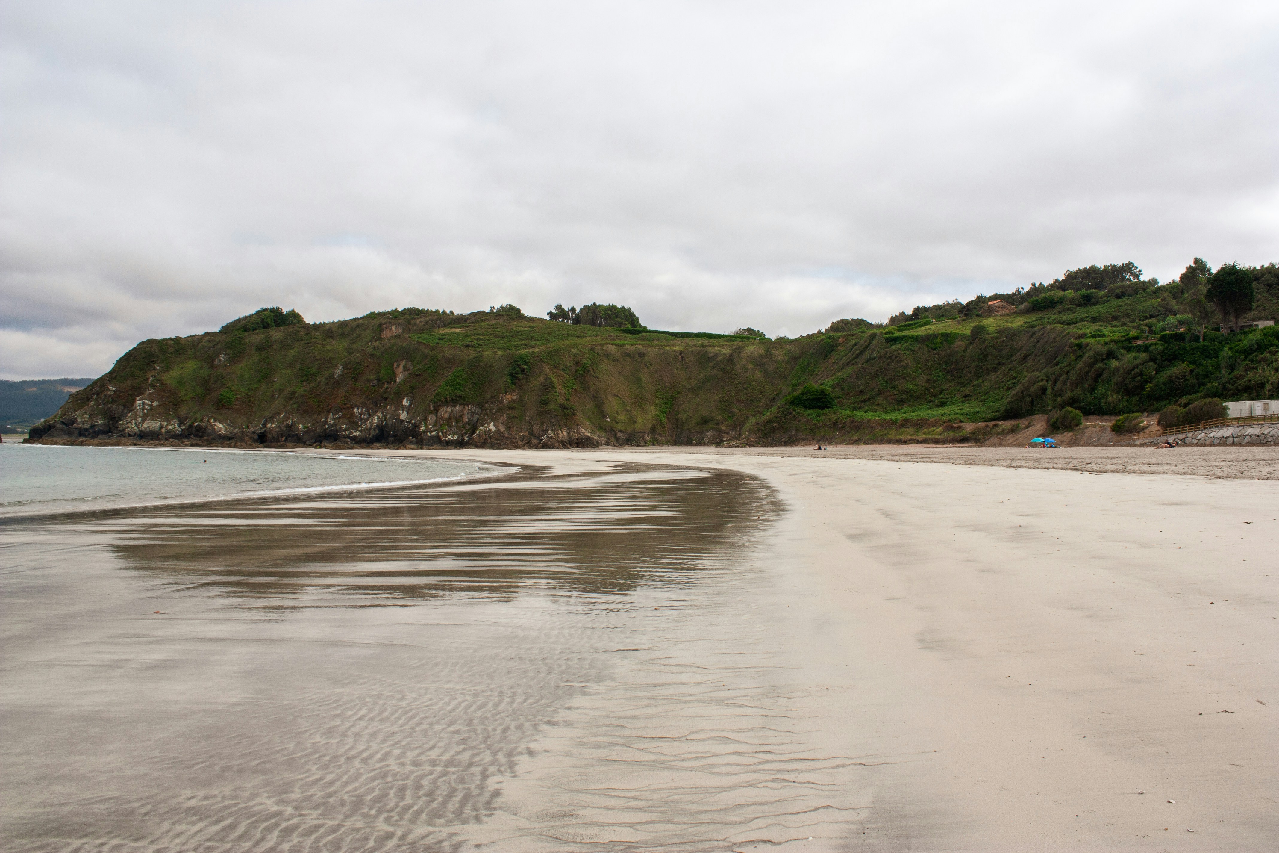 A wide sandy beach with wet sand reflecting a cloudy sky below green hills