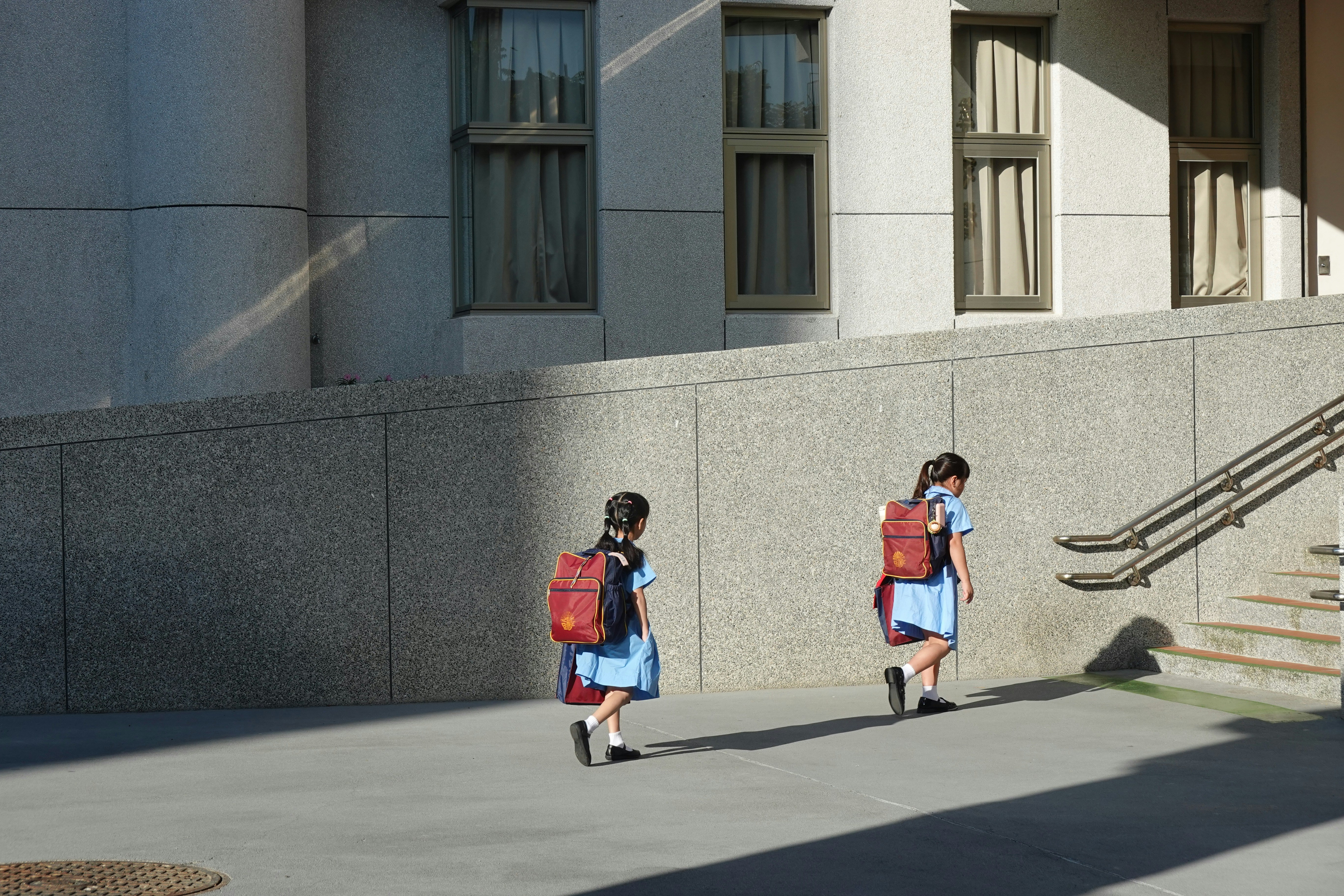 Deux écolières en uniforme bleu, vues de dos, marchent avec leur cartable le long d'un bâtiment