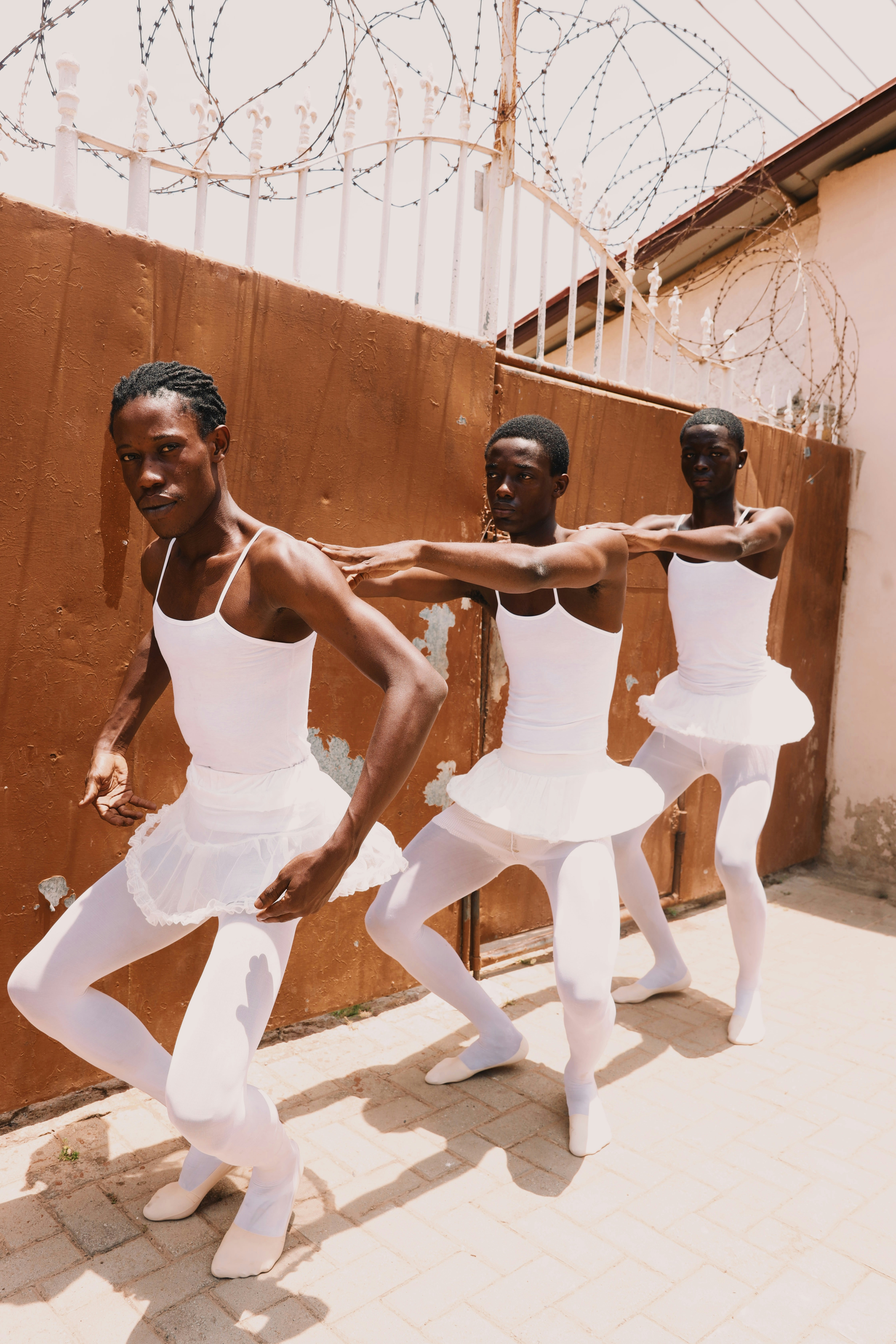 Three men in white leotards and tutus dancing before a brown metal gate