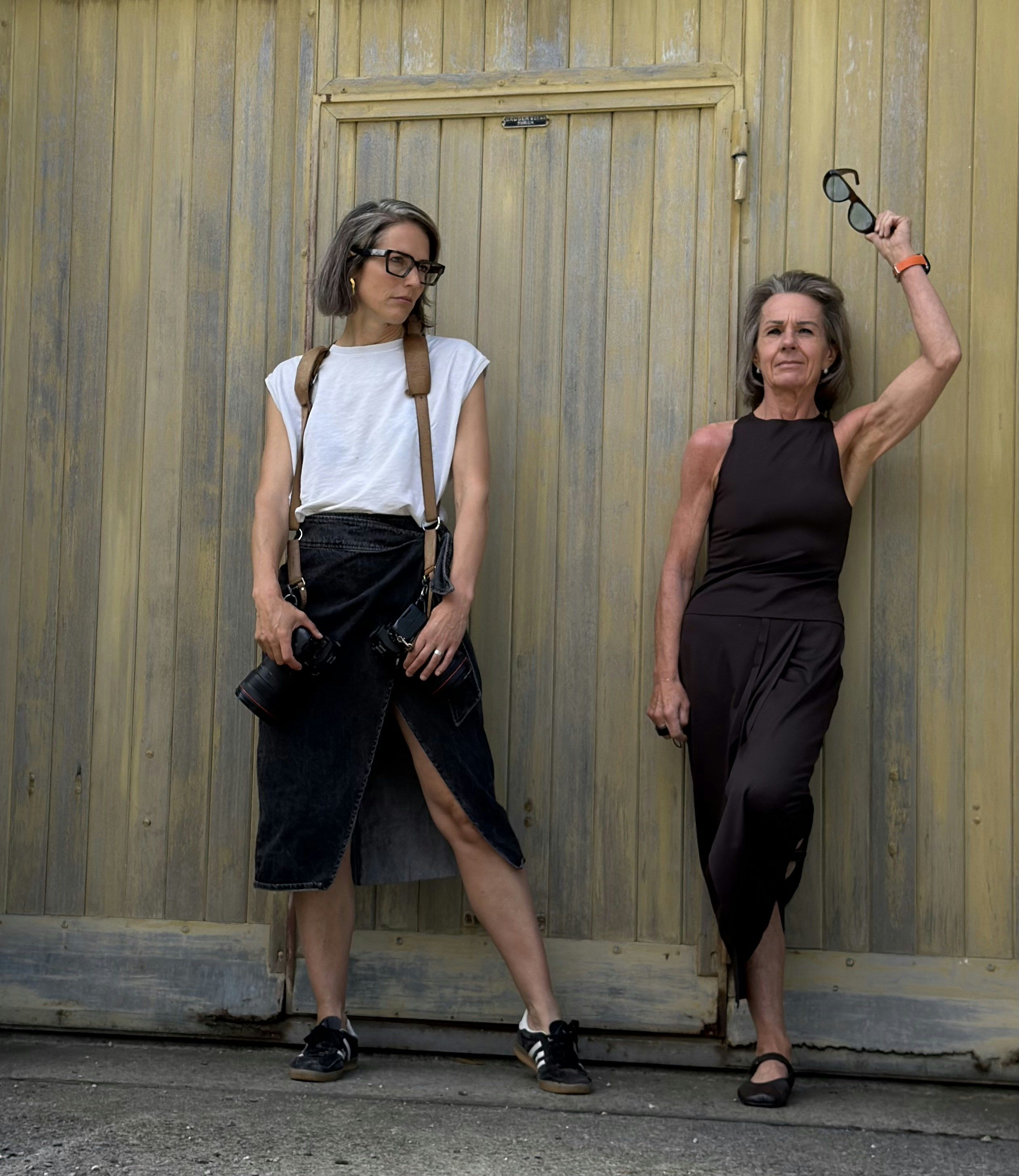 Two women in dark clothing against a weathered yellow wooden door