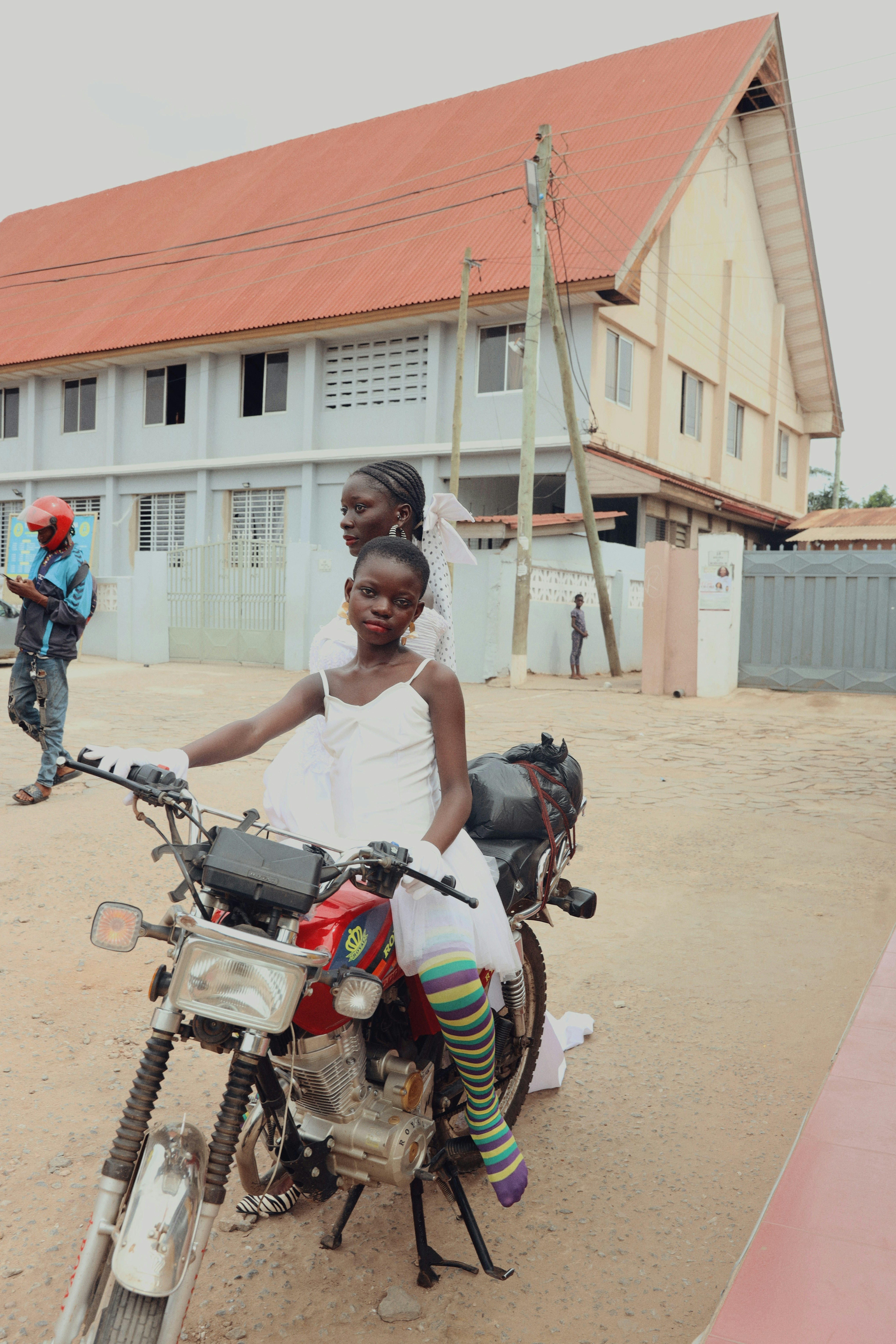 Two people in white dresses on a red motorcycle in a paved courtyard