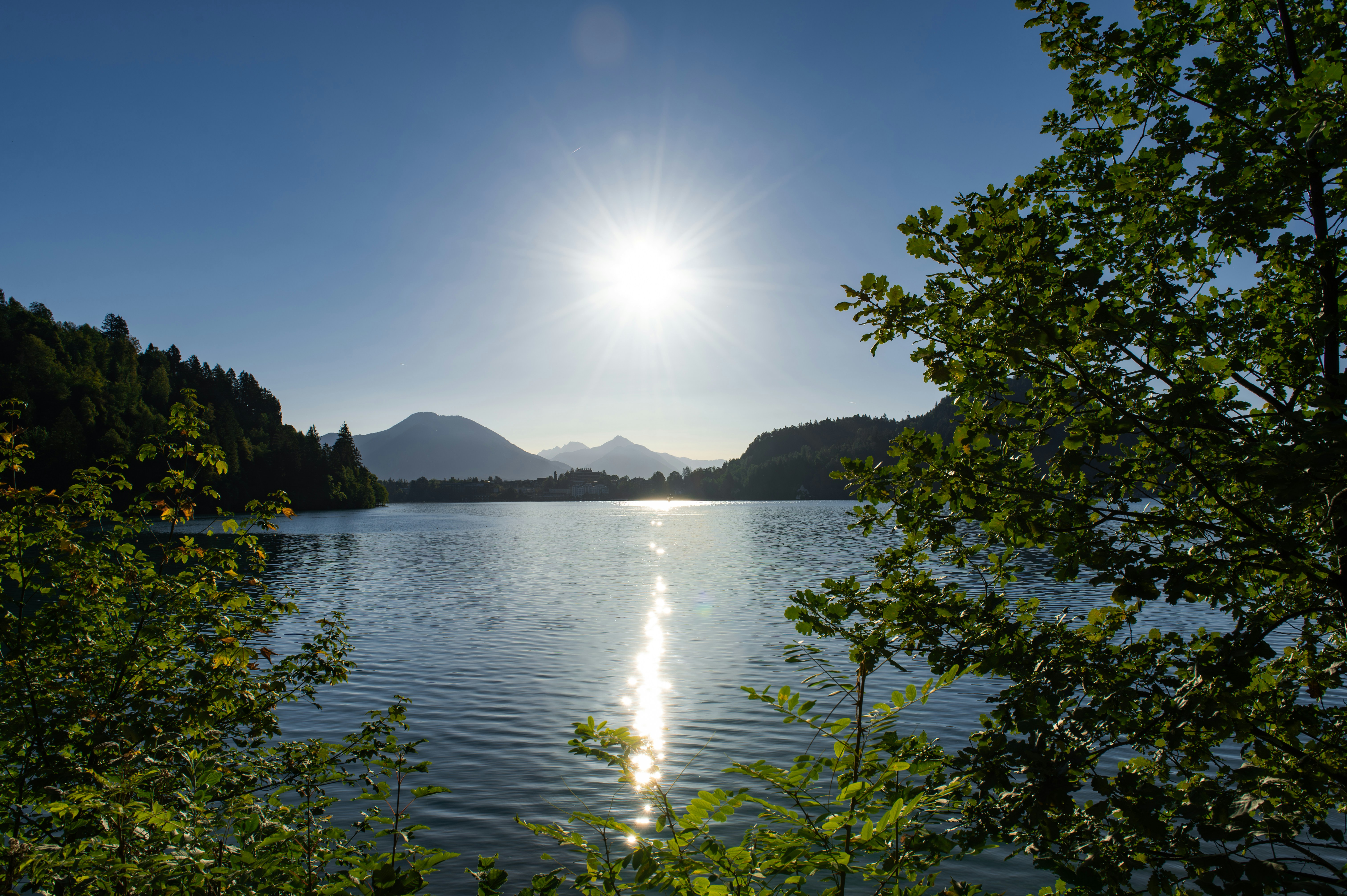A bright sun over a calm lake framed by green trees and distant mountains