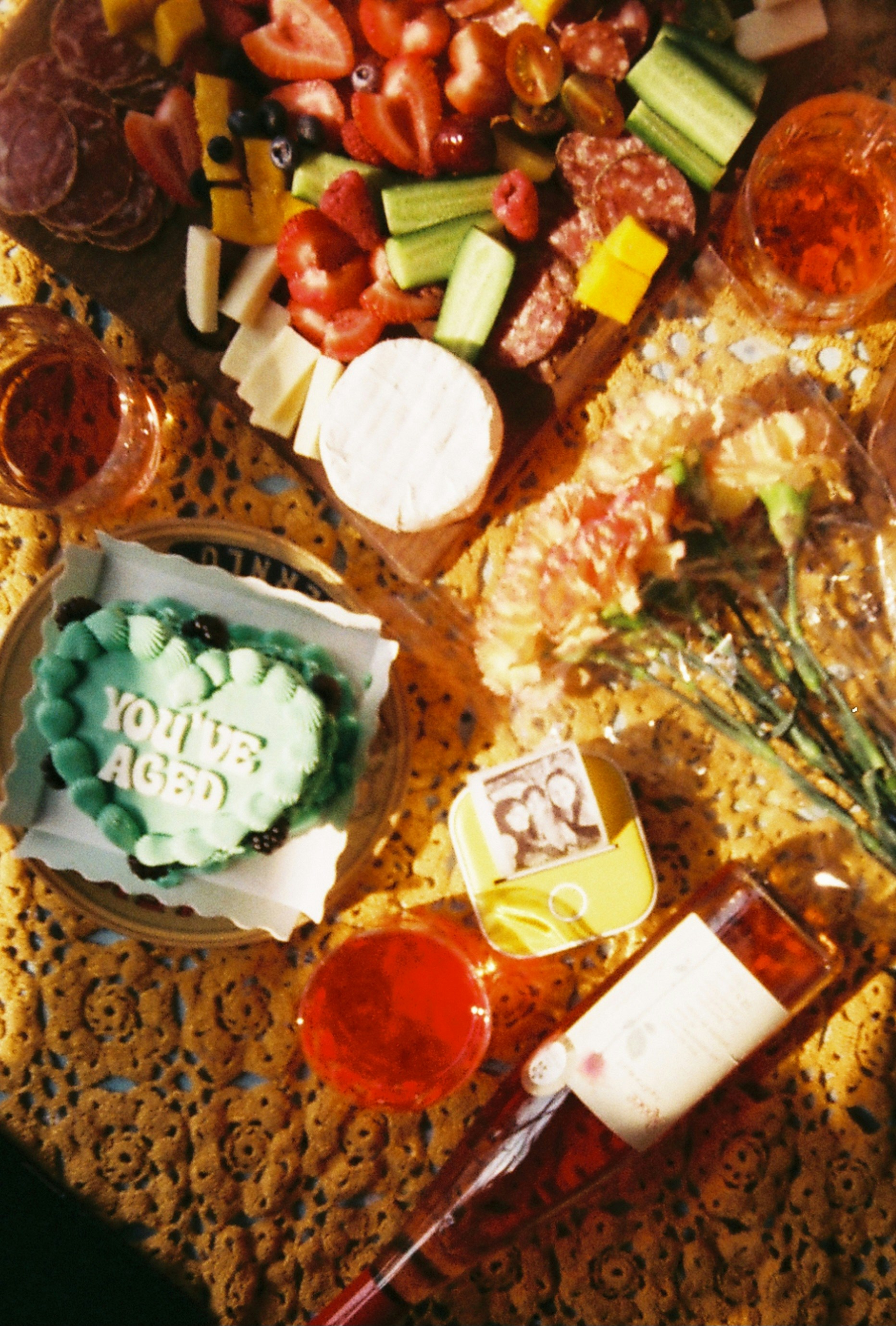 A heart-shaped cake reading YOU'VE AGED, charcuterie board, wine, and flowers on lace