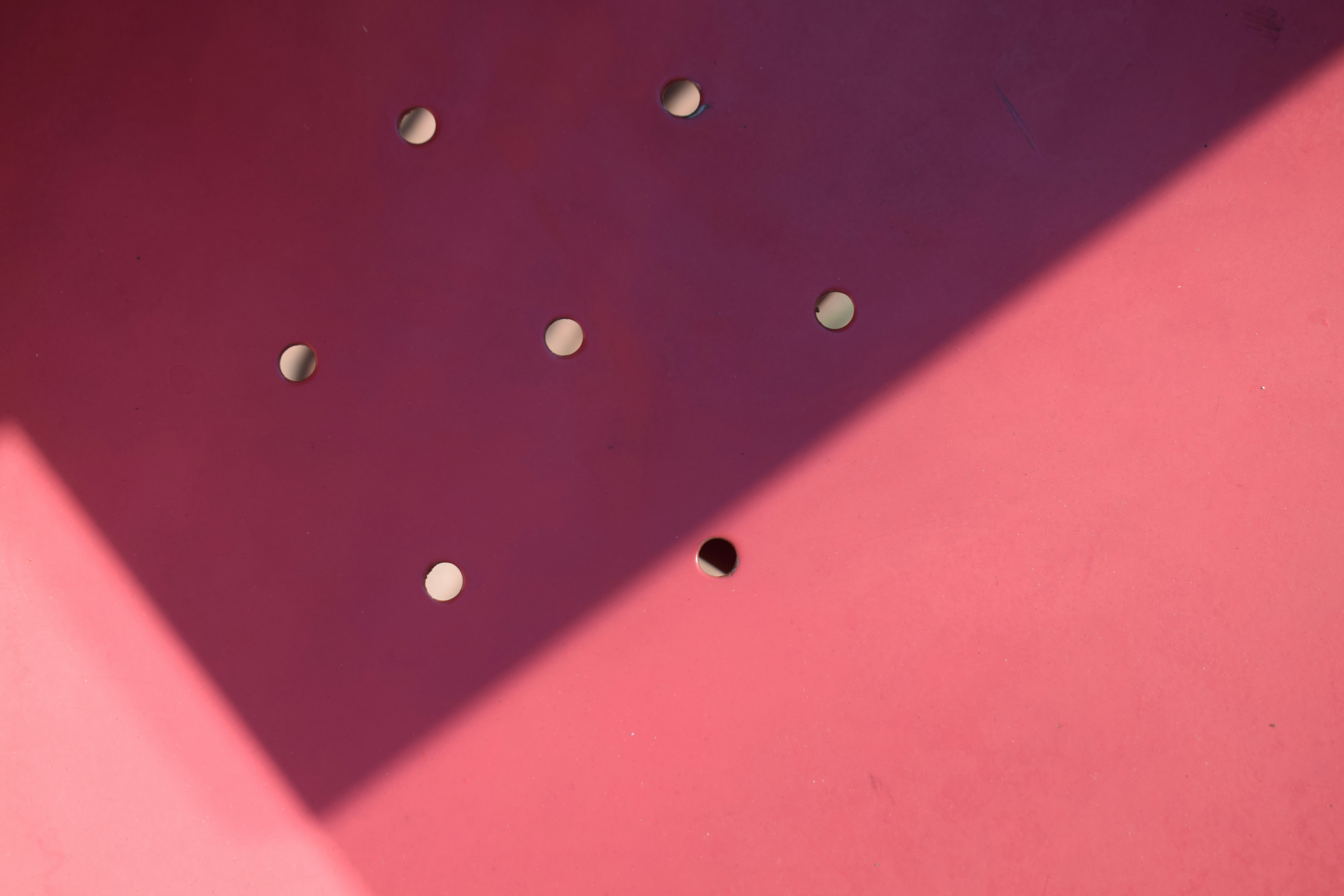 A pink metal surface with circular holes and a sharp diagonal shadow