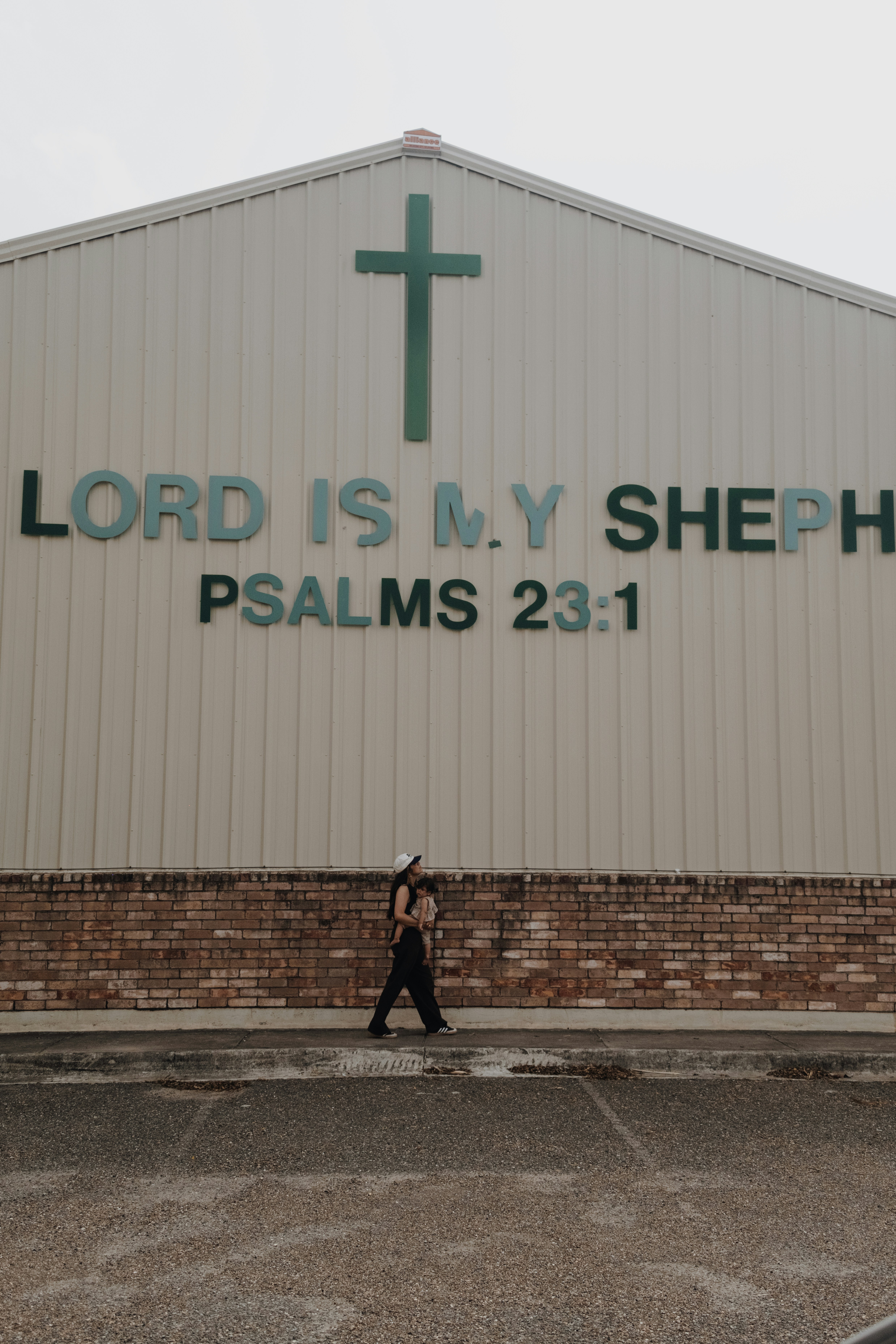 A person carrying a child walking past a building with a cross and text