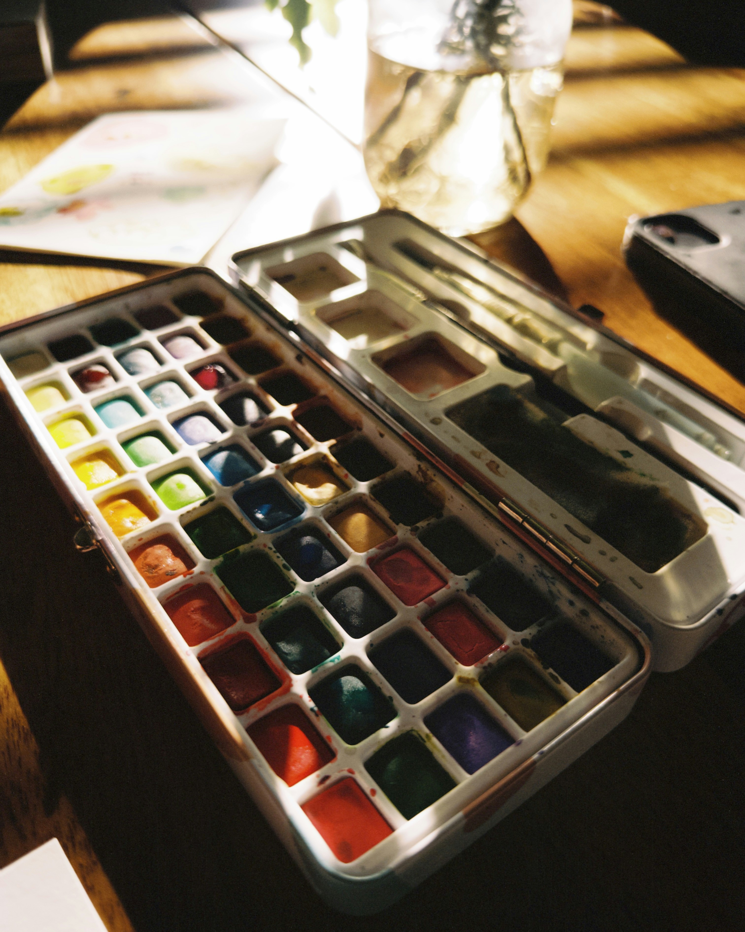 A metal watercolor paint set on a wooden table in warm sunlight