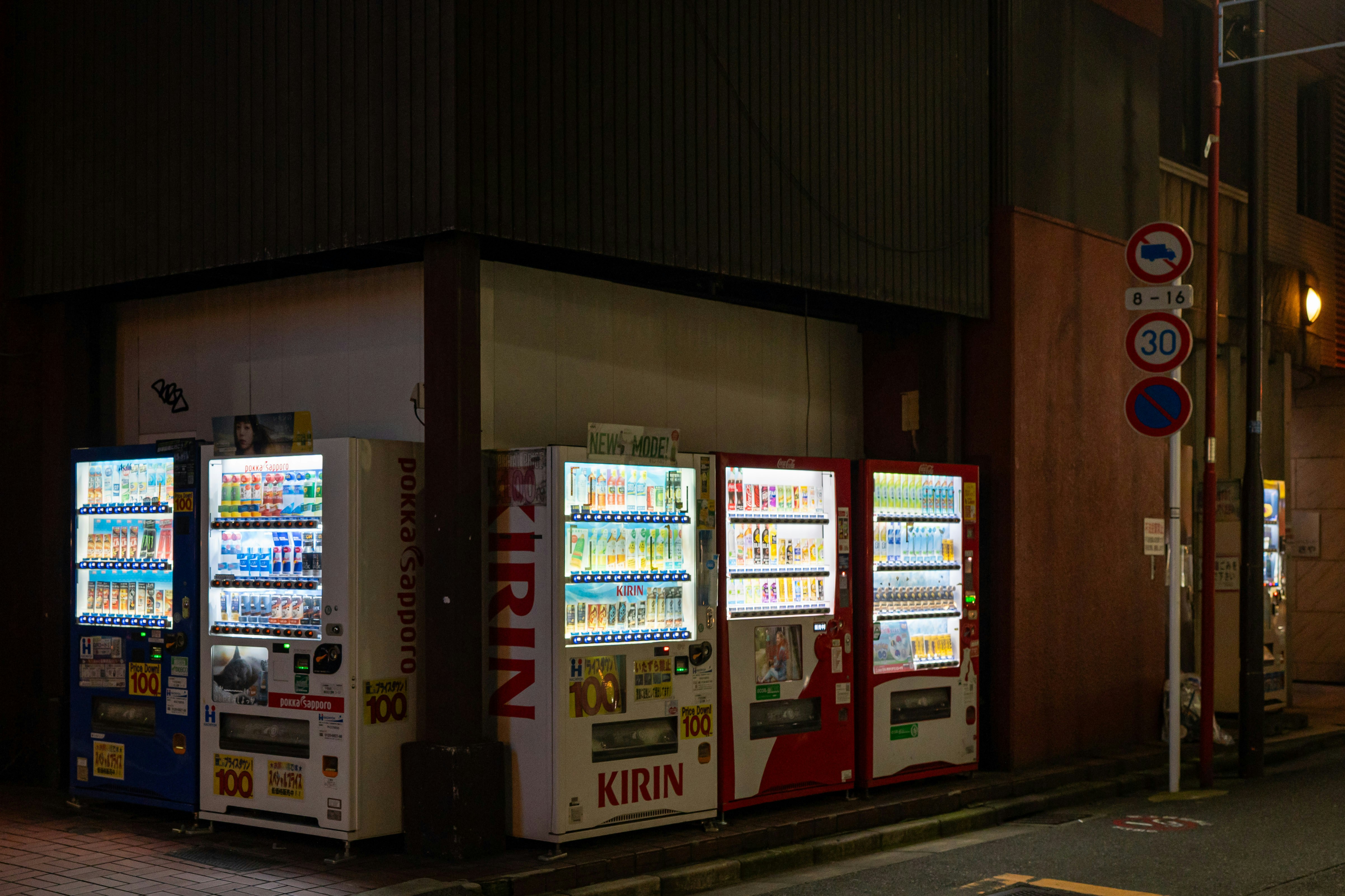 夜の暗い建物の壁際に立つ4台の点灯した自動販売機