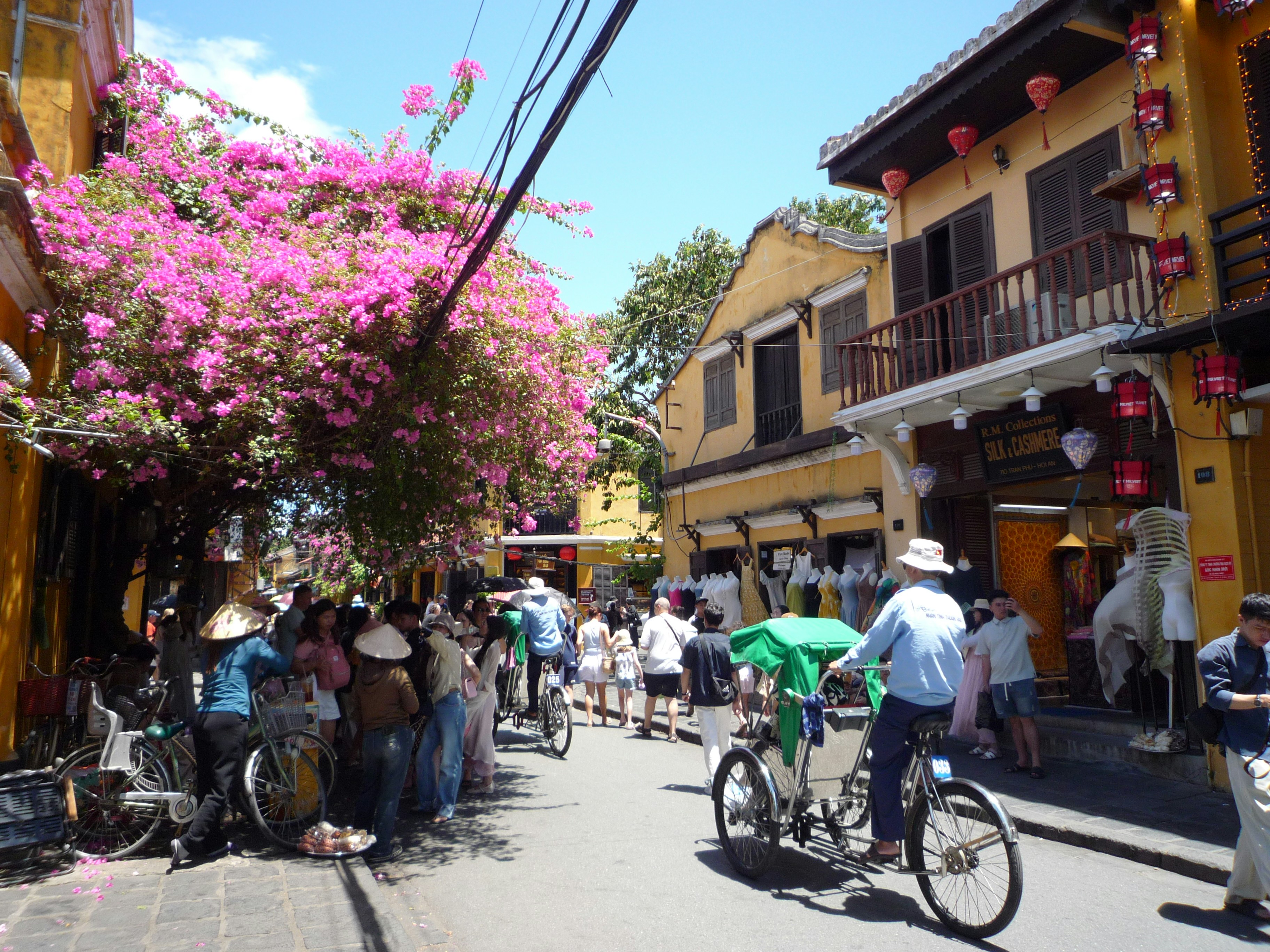 A busy street in Hoi An with yellow buildings, pink bougainvillea, and a pedicab