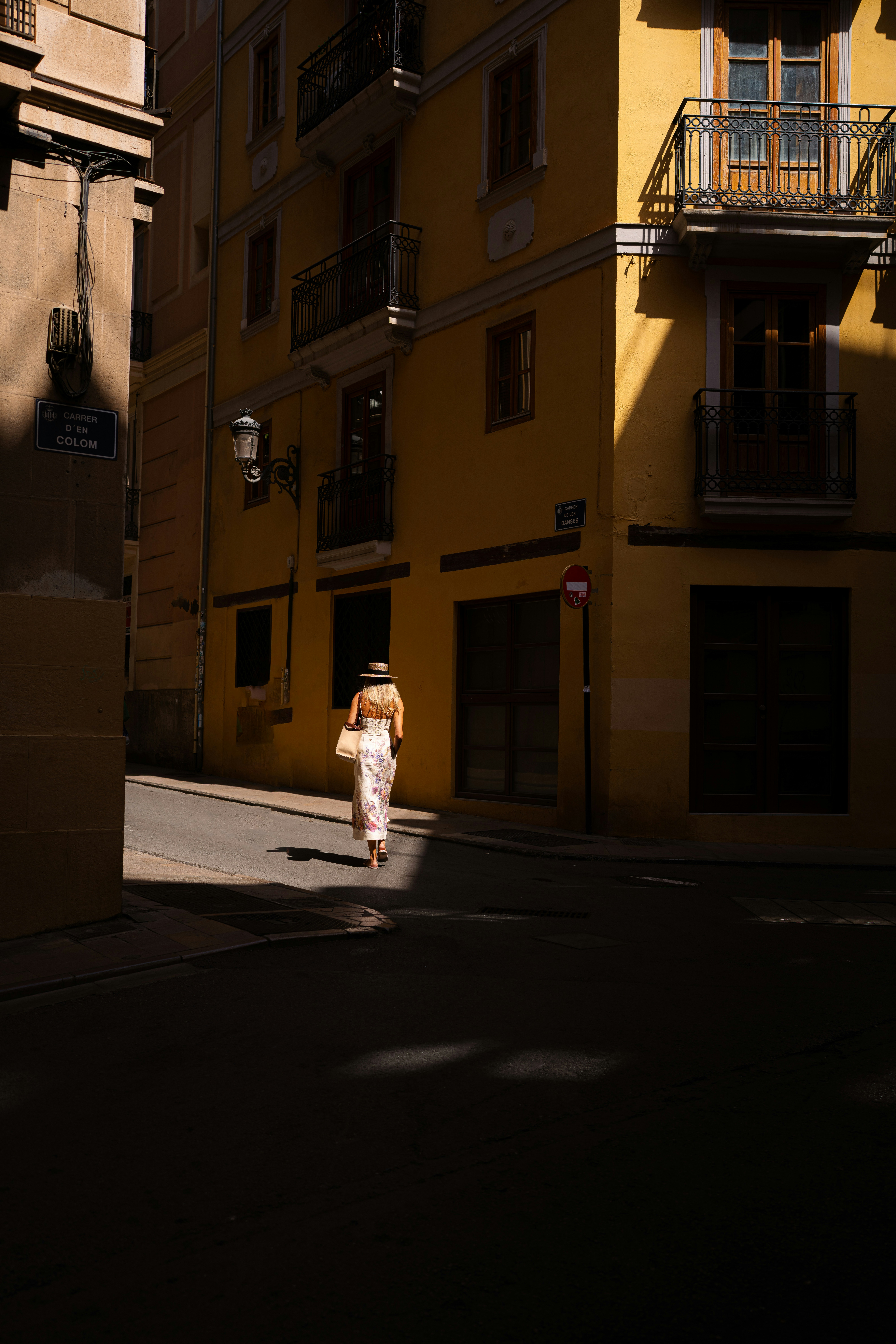 A woman in a floral dress and hat walking down a sunlit yellow street