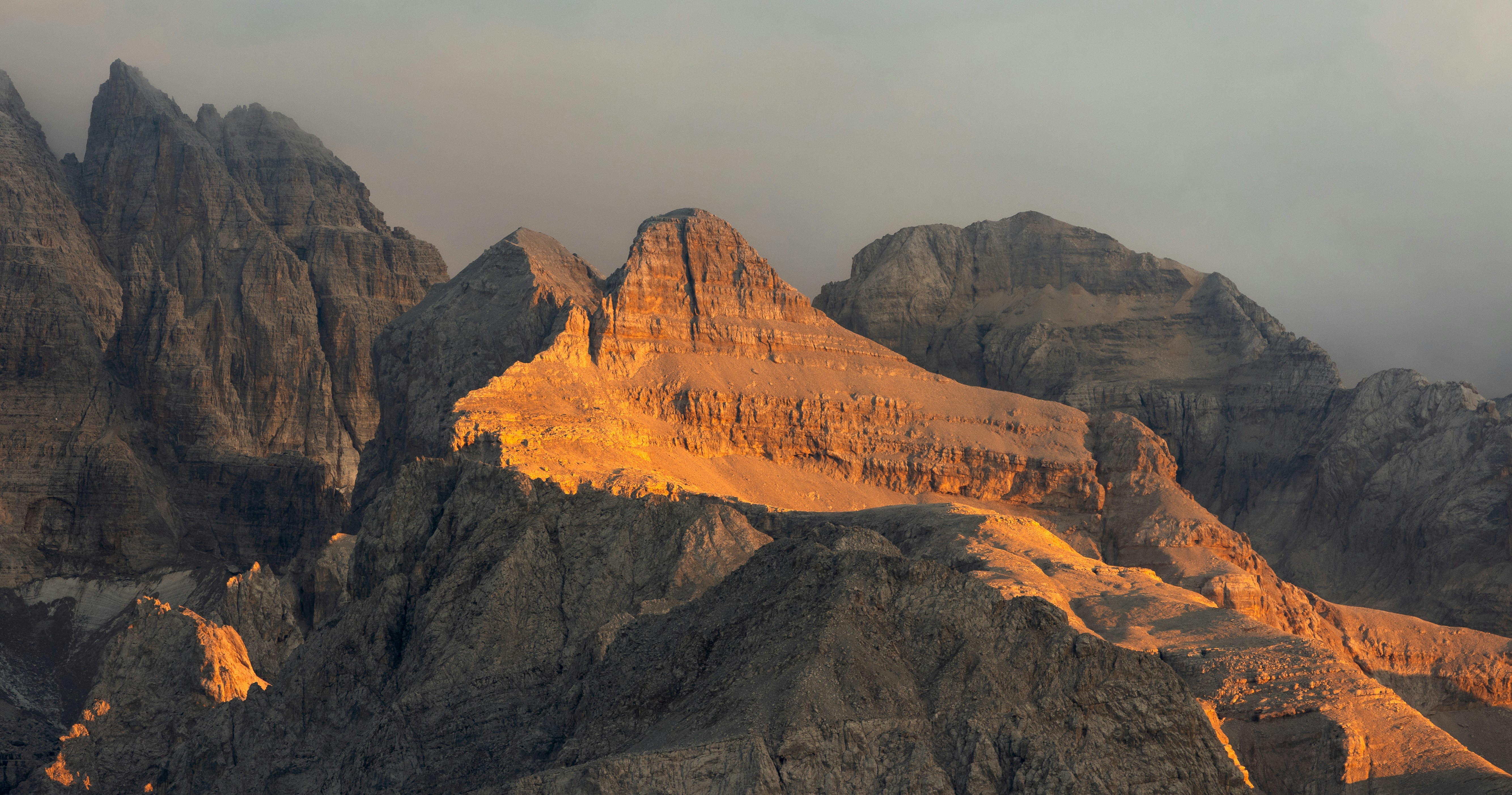 Jagged rocky mountain peaks illuminated by warm golden light against a grey sky