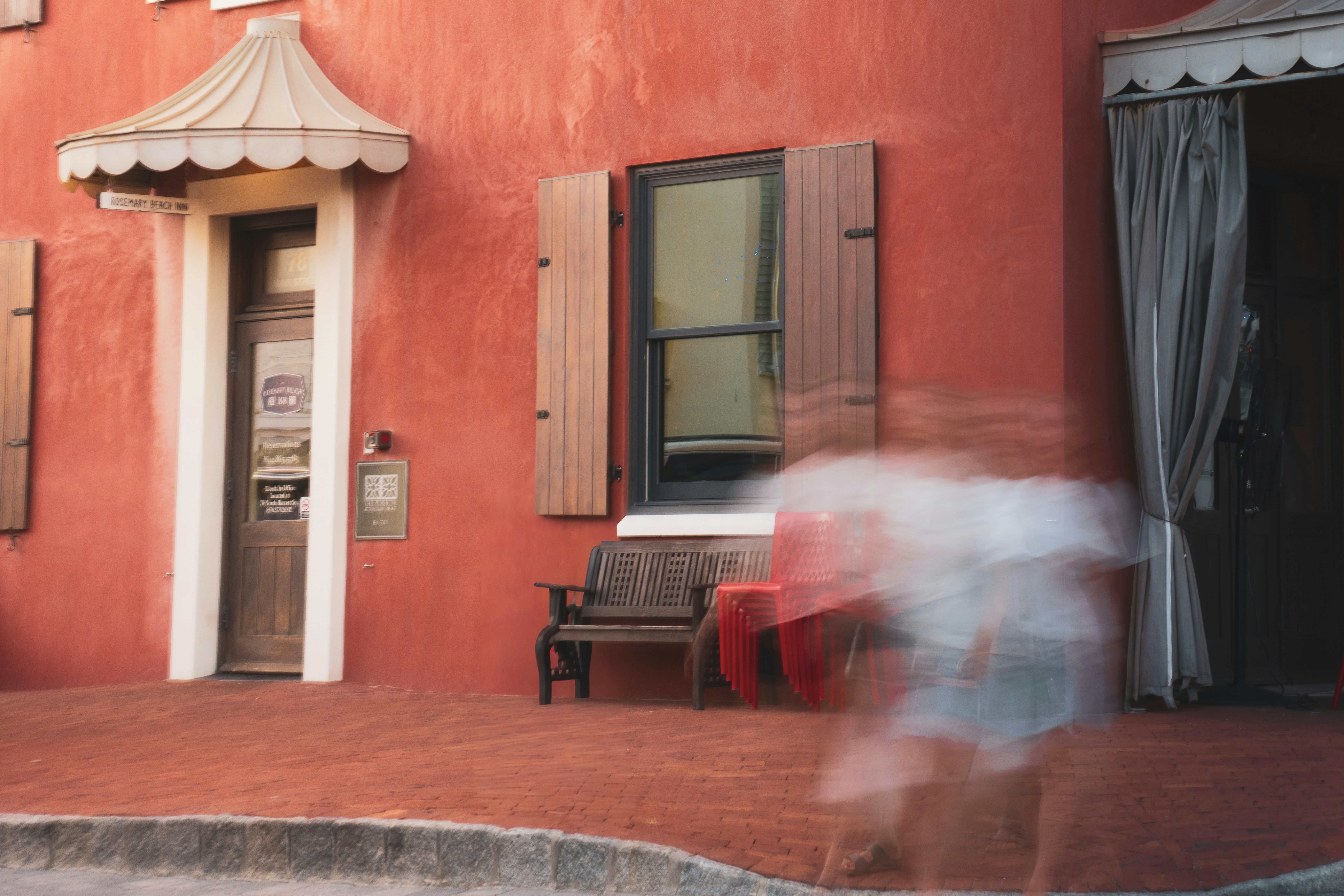 A blurred person walking past a red building with a wooden bench