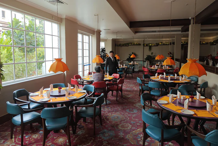 A restaurant room with round tables, teal chairs and orange pendant lights.