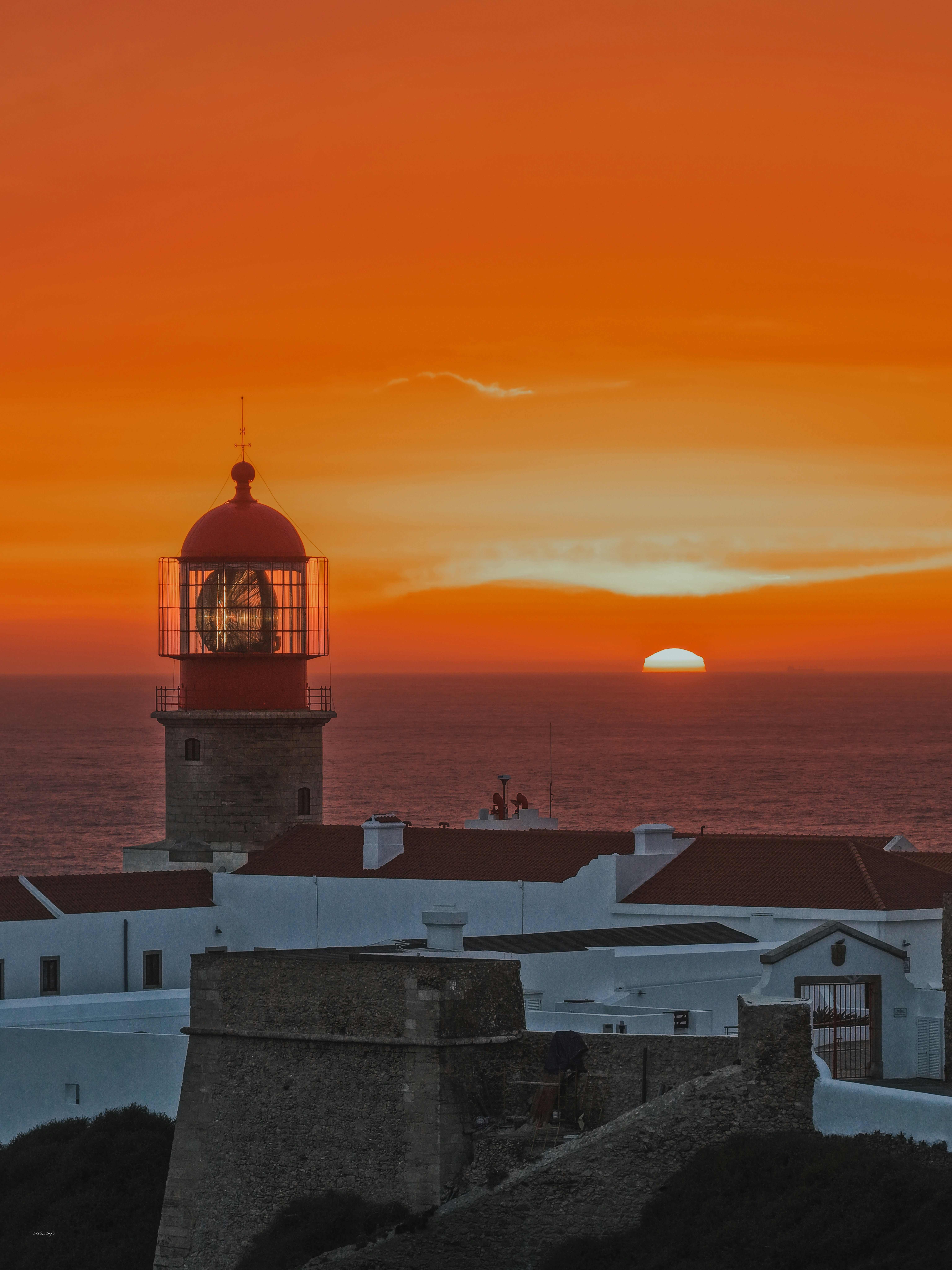 A lighthouse with a red lantern room above white buildings at low sun