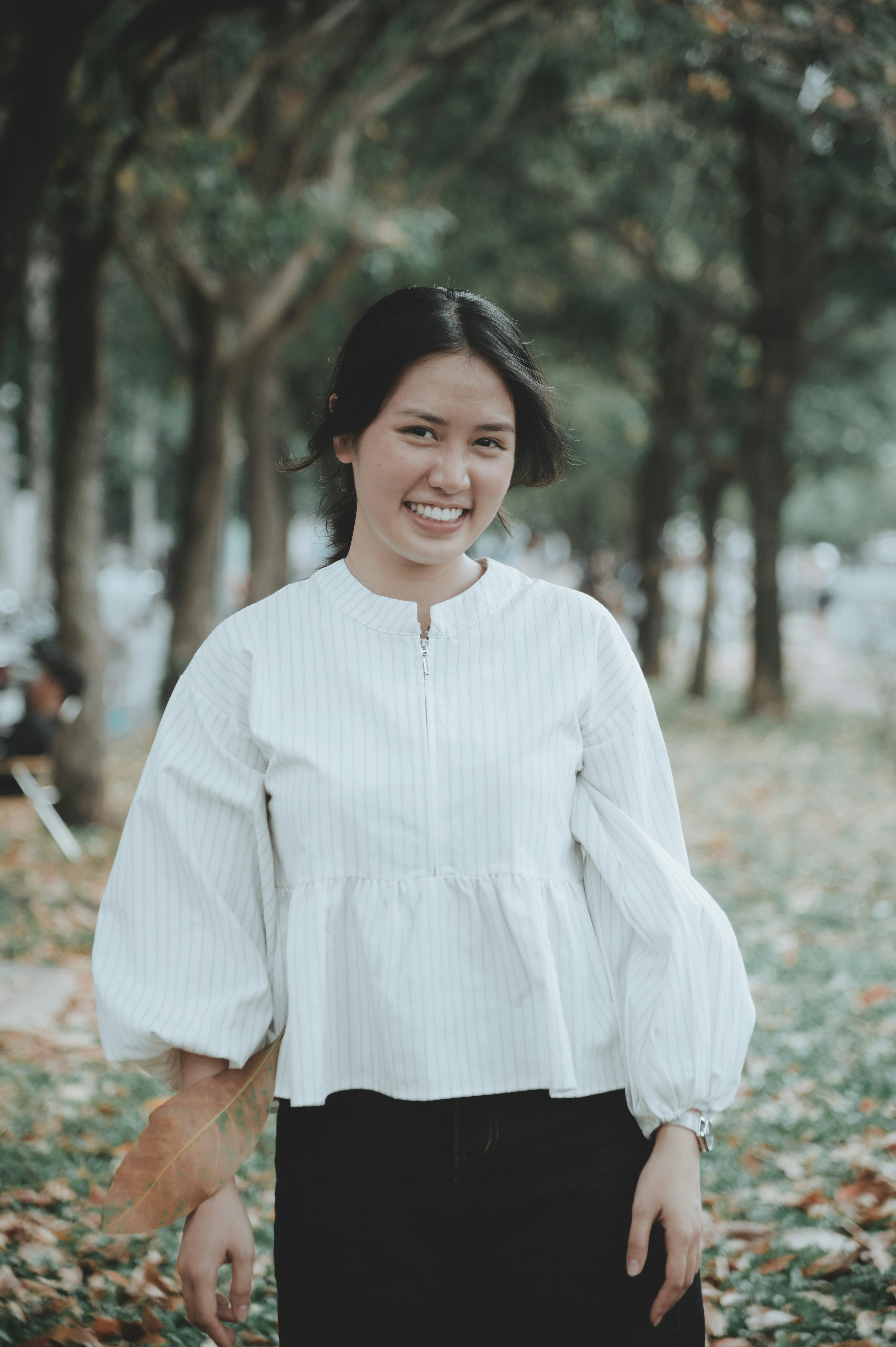 Una mujer sonriente con una blusa blanca de rayas sosteniendo una hoja caída al aire libre