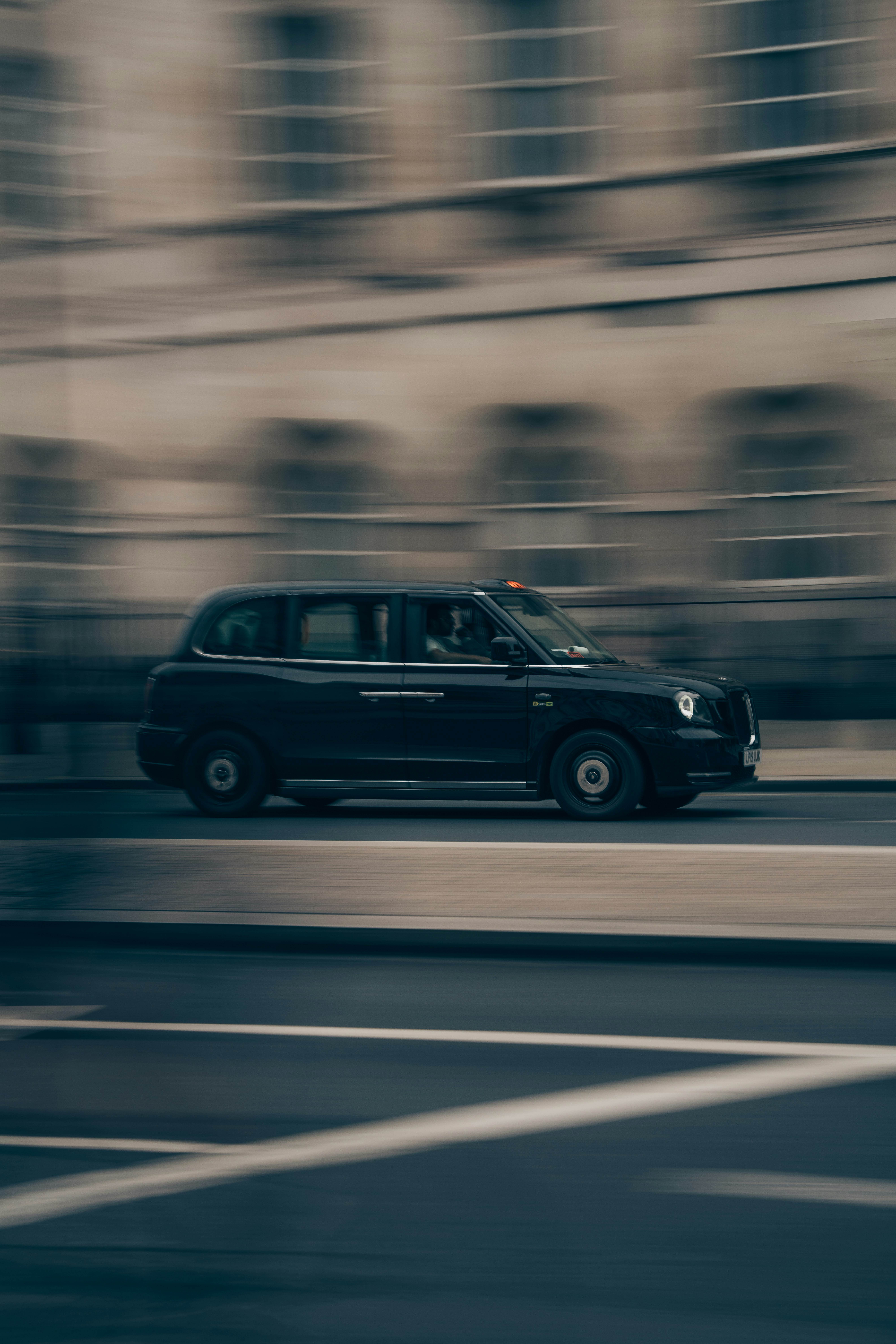 A black London taxi driving past a blurred building in a city street