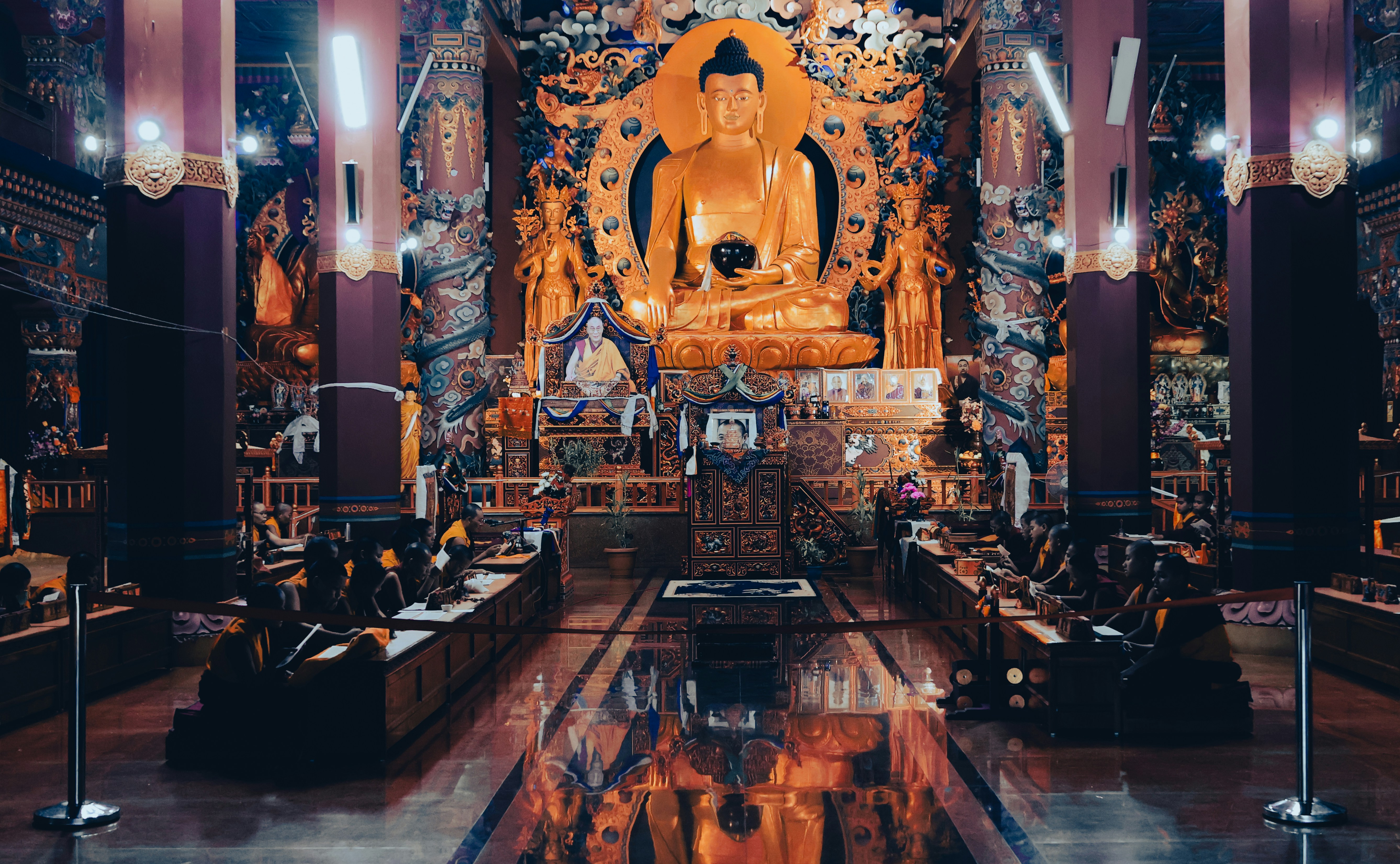 Monks in robes sitting at desks inside a temple with a large golden Buddha statue
