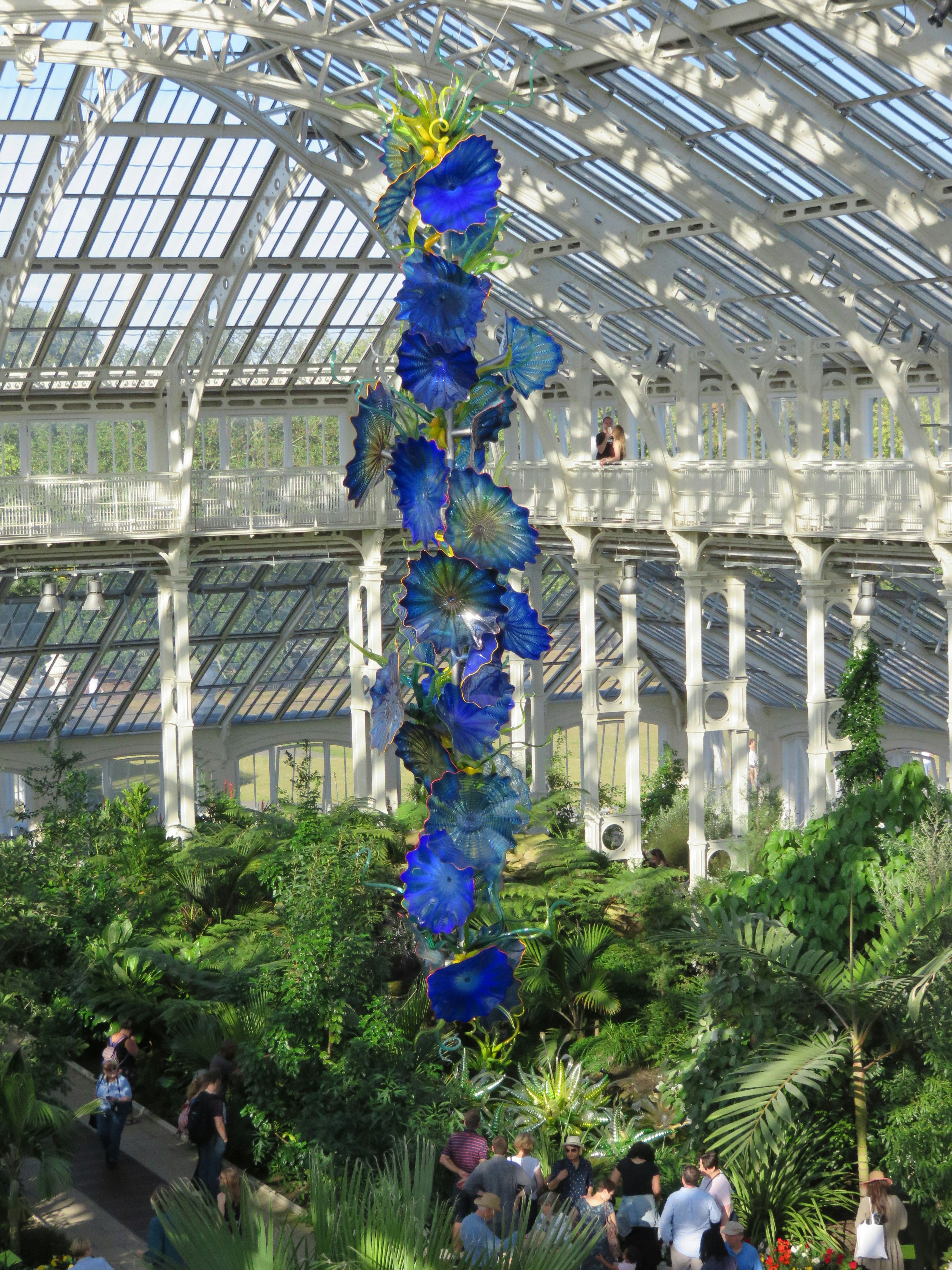 A tall blue glass flower sculpture rises within a glasshouse, surrounded by lush greenery and visitors on a balcony above. The scene emphasizes the botanical display and architectural context.