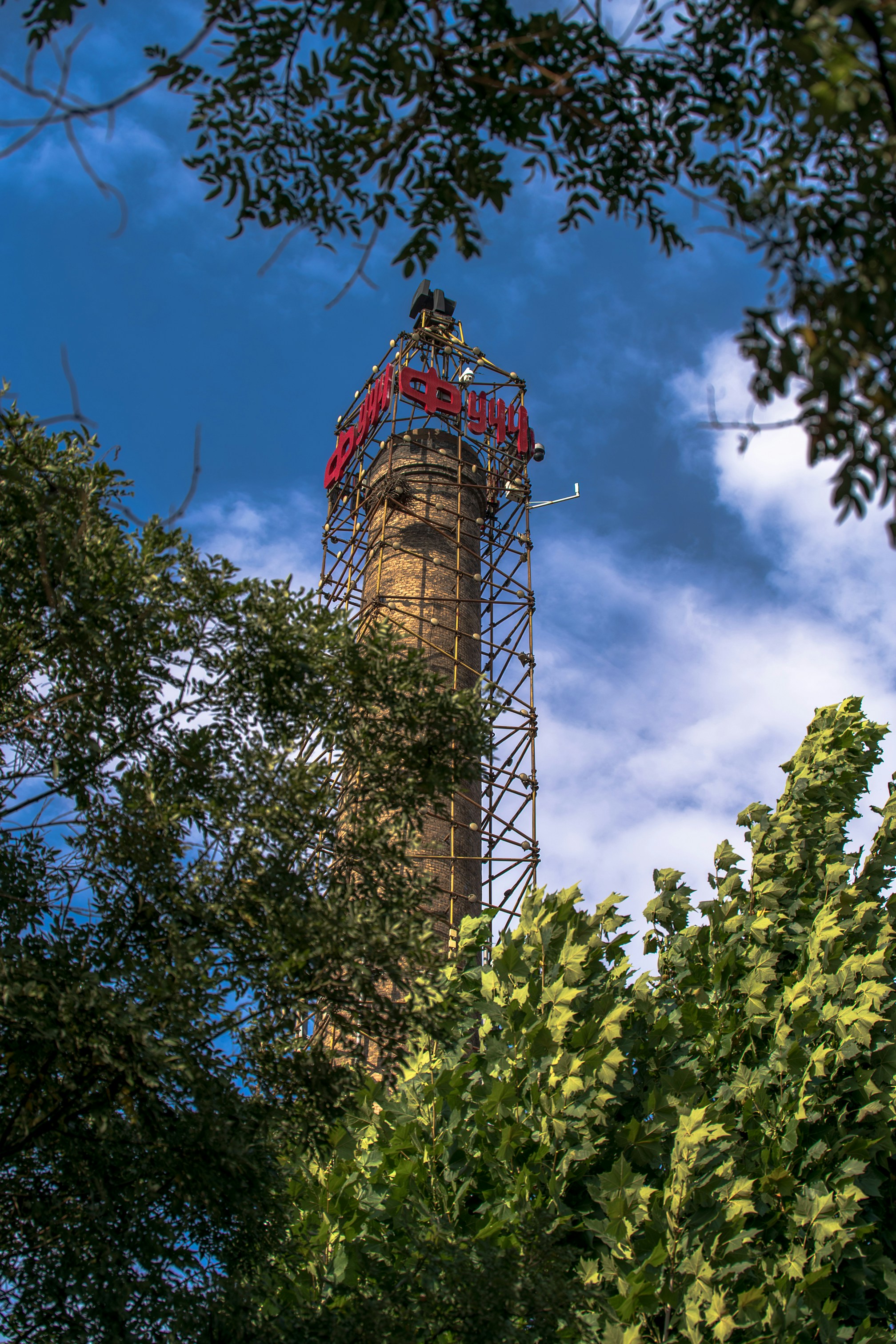 A tall brick industrial chimney surrounded by green trees under a blue sky