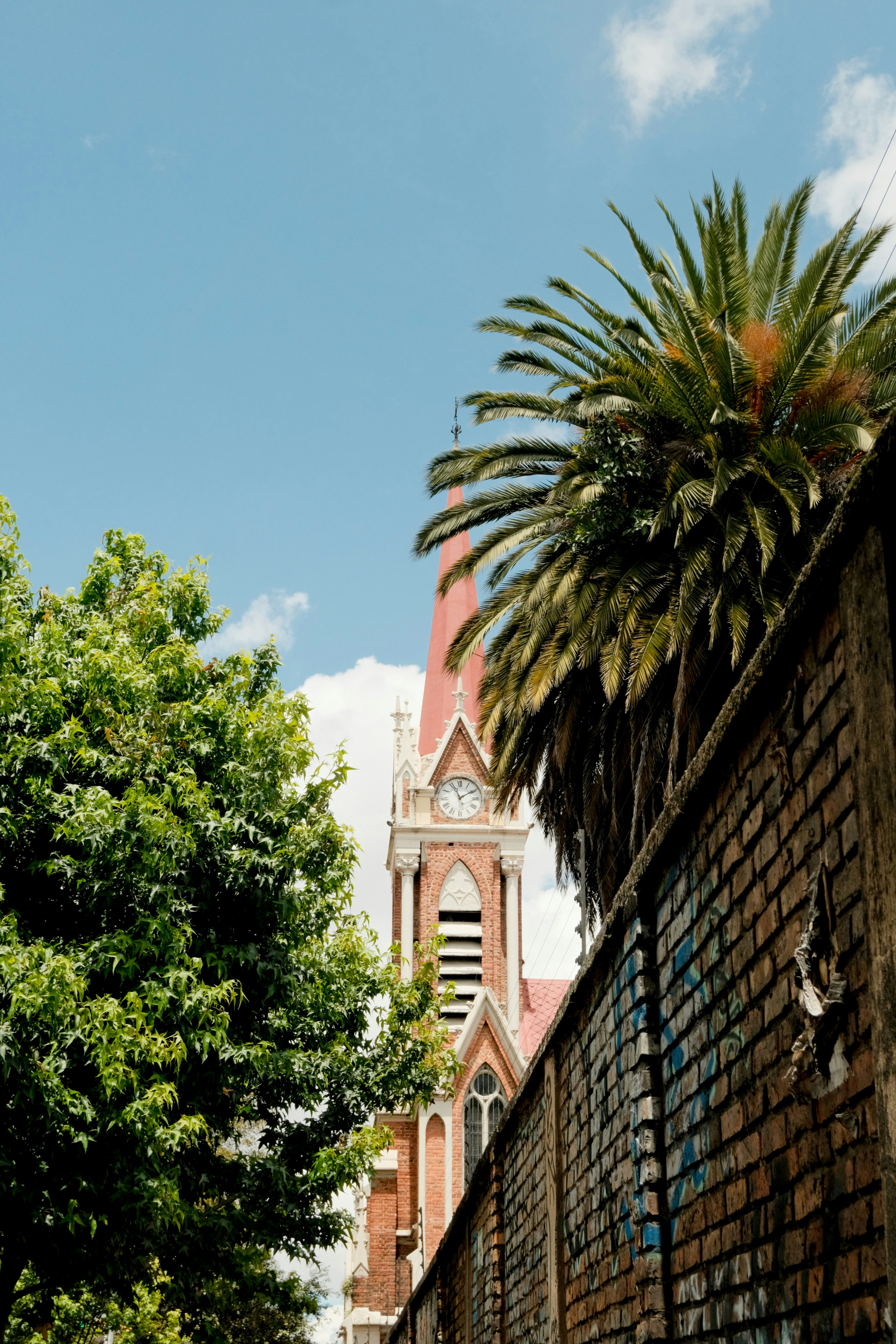 A red-roofed church tower behind a brick wall and lush green trees