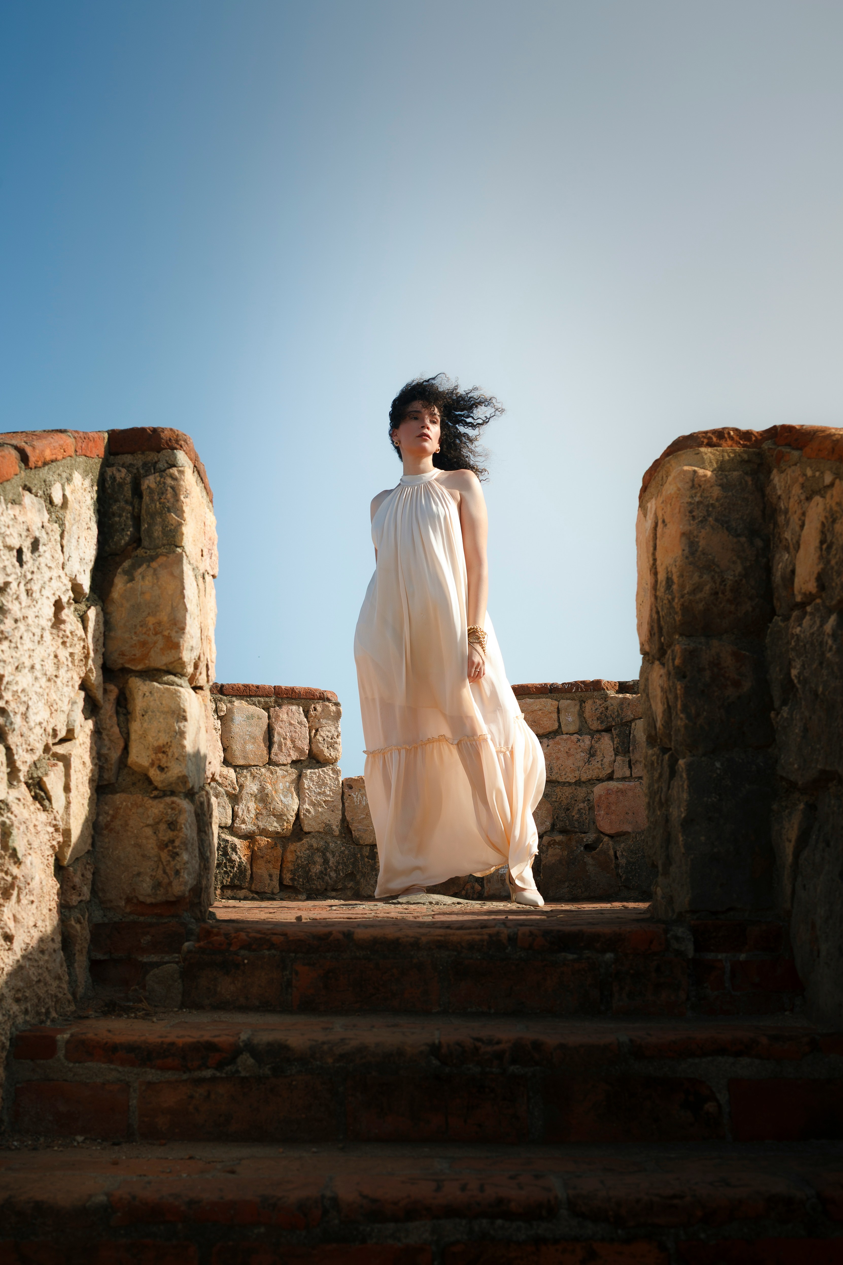 A woman in a flowing cream dress on stone steps between old walls