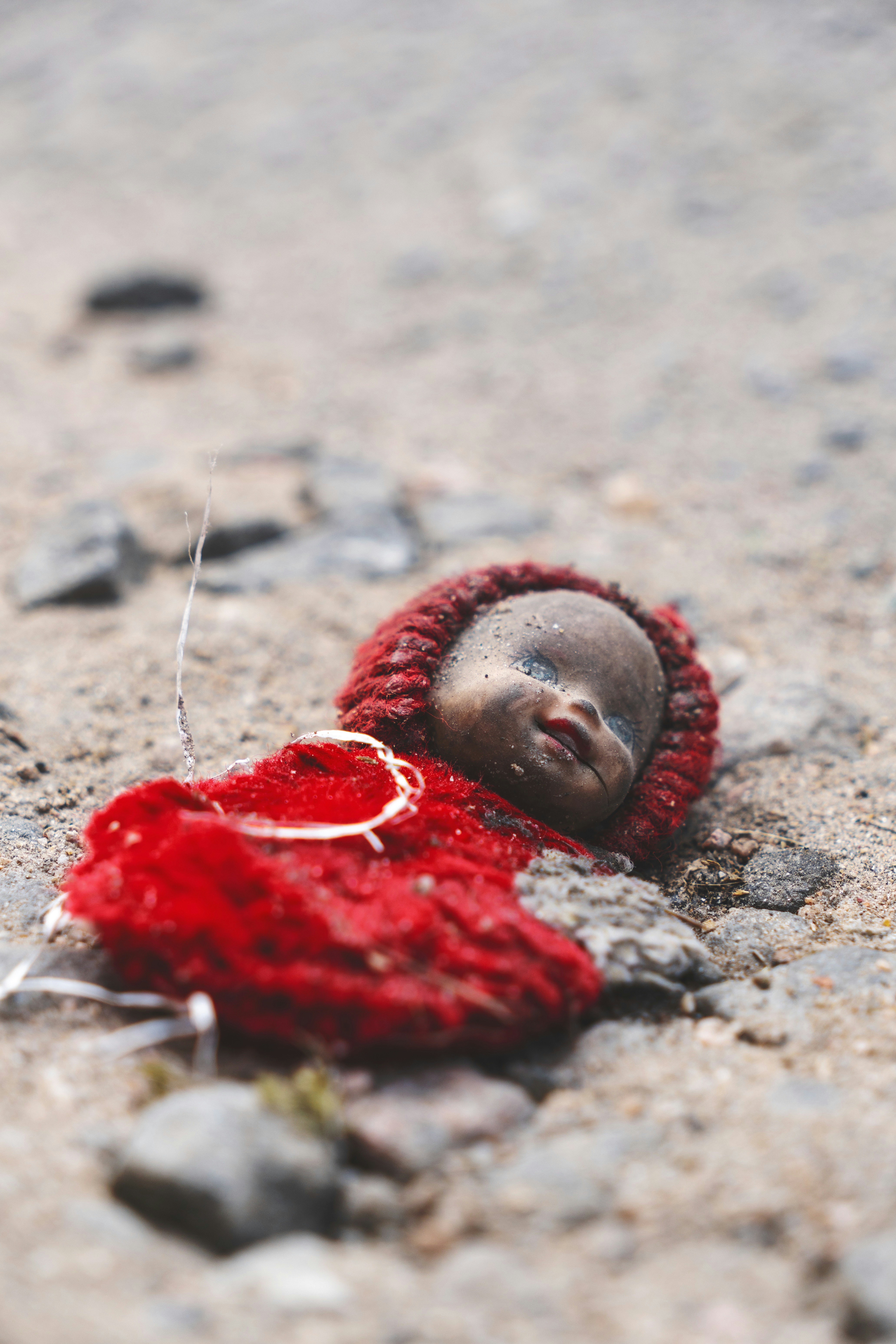 A dirty doll in a red knitted outfit lying on gravel ground