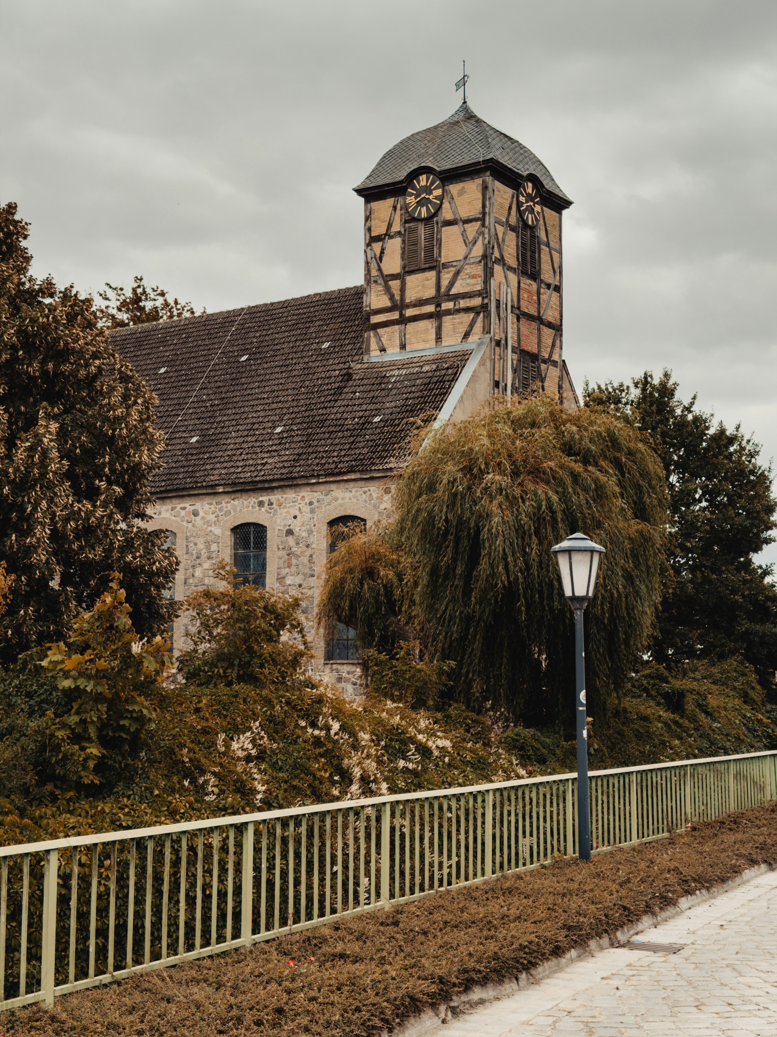 A stone church with a timber-framed clock tower behind trees and a metal fence