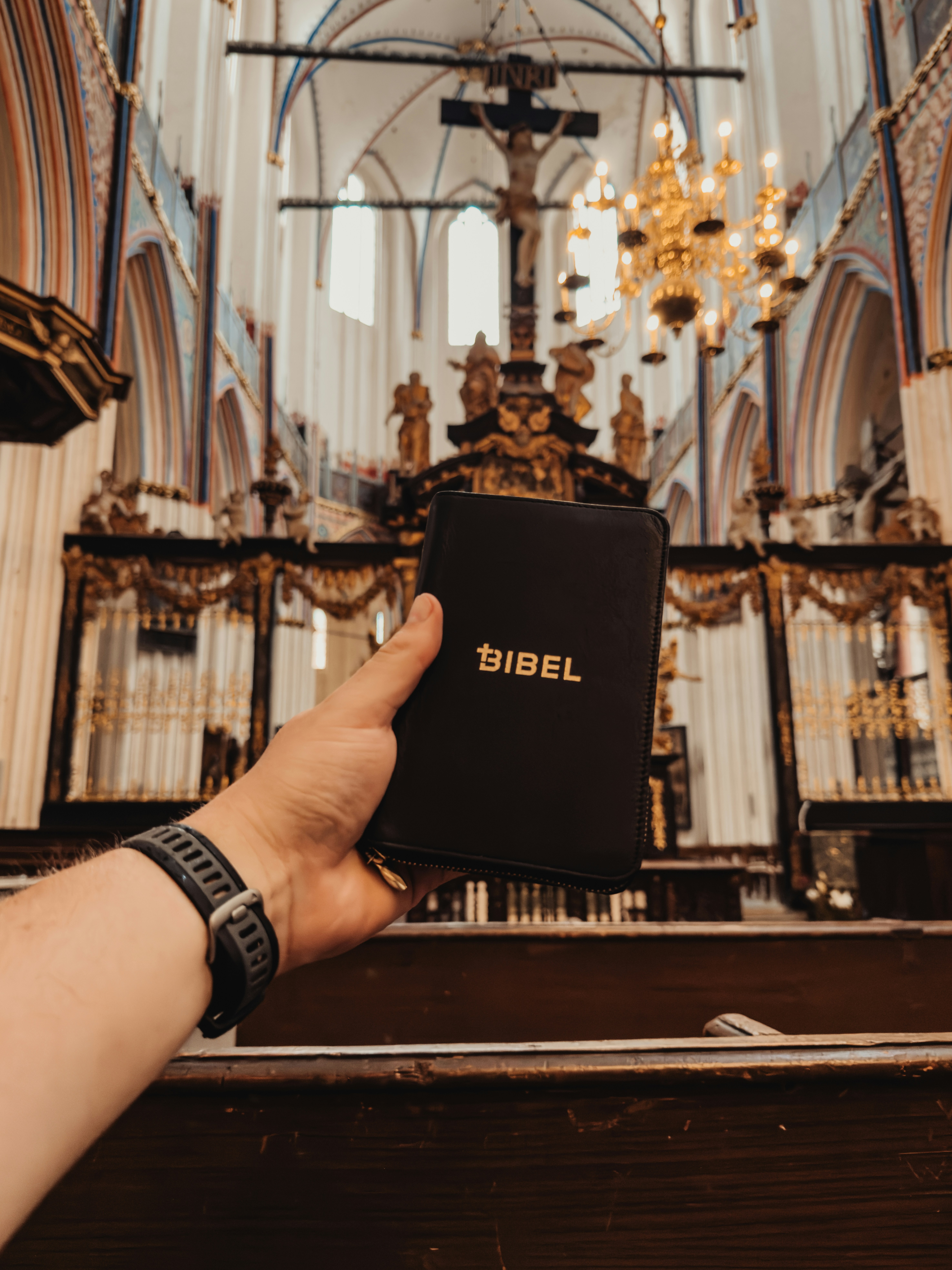 A hand holding a black Bible in a church with a crucifix and chandelier