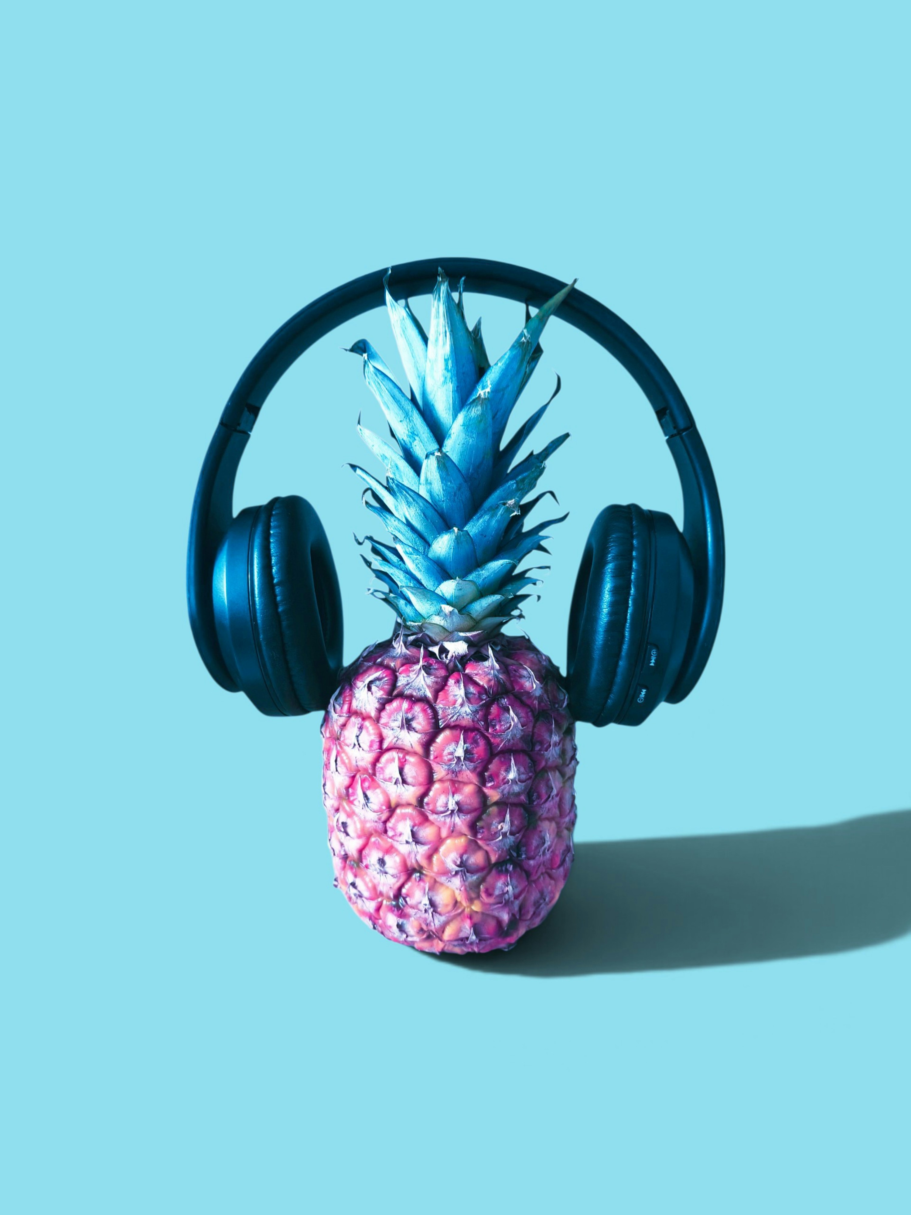 A pineapple wearing black over-ear headphones against a solid light blue background