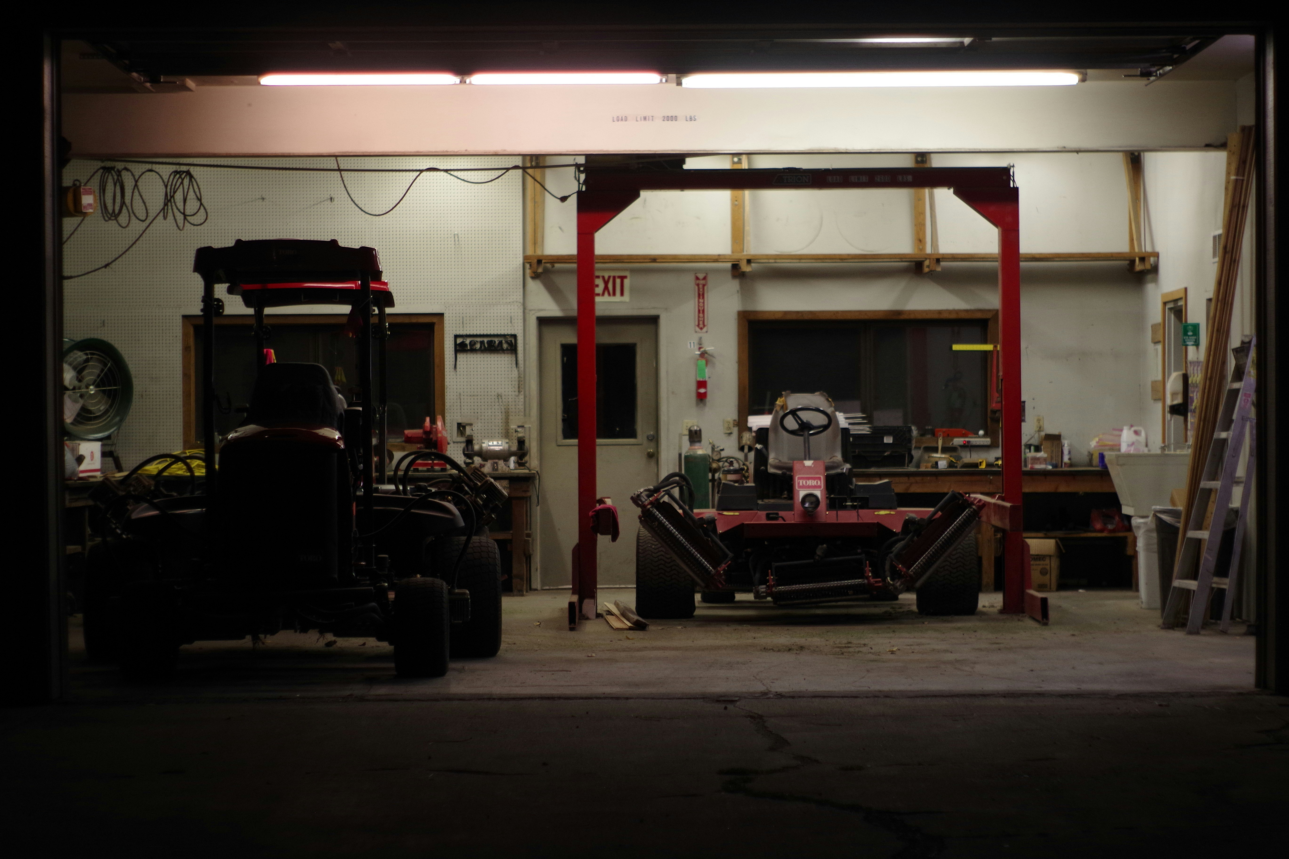 Two red industrial lawn mowers inside a dimly lit maintenance garage with a red metal lift