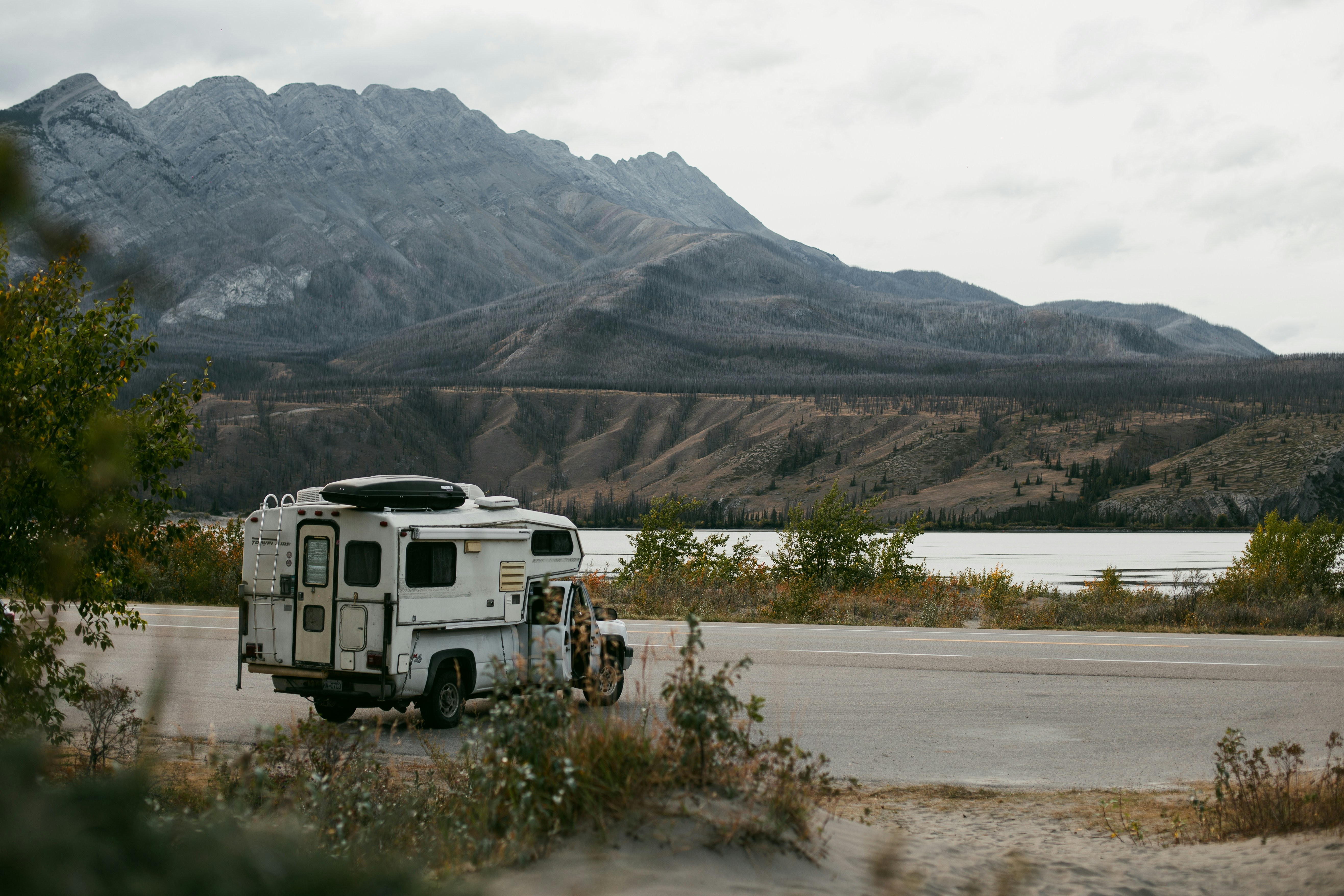 Camping-car à cellule garé au bord d'une route, devant un lac et des montagnes sous un ciel couvert