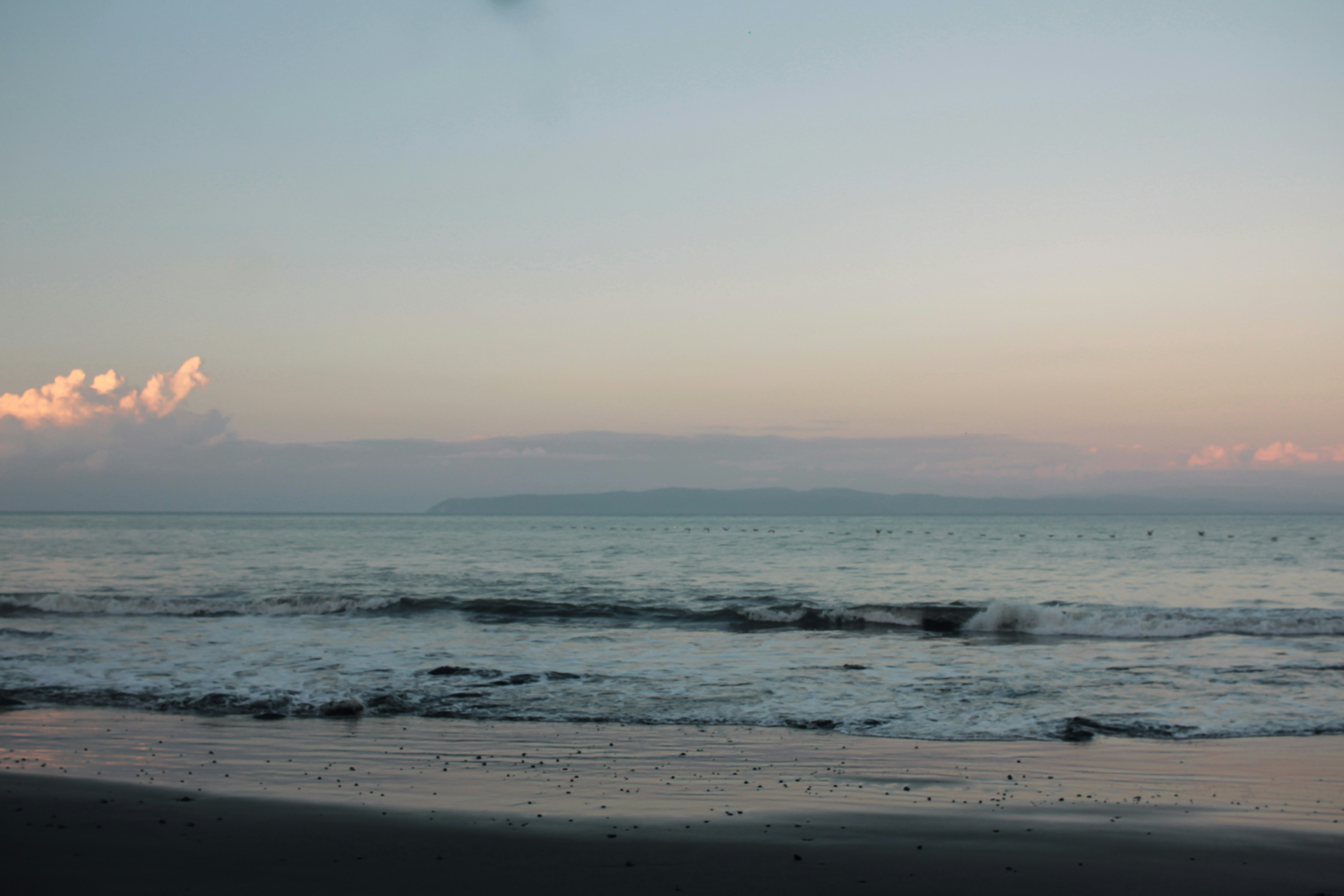 Gentle waves rolling onto a dark sandy beach under a soft pastel sky