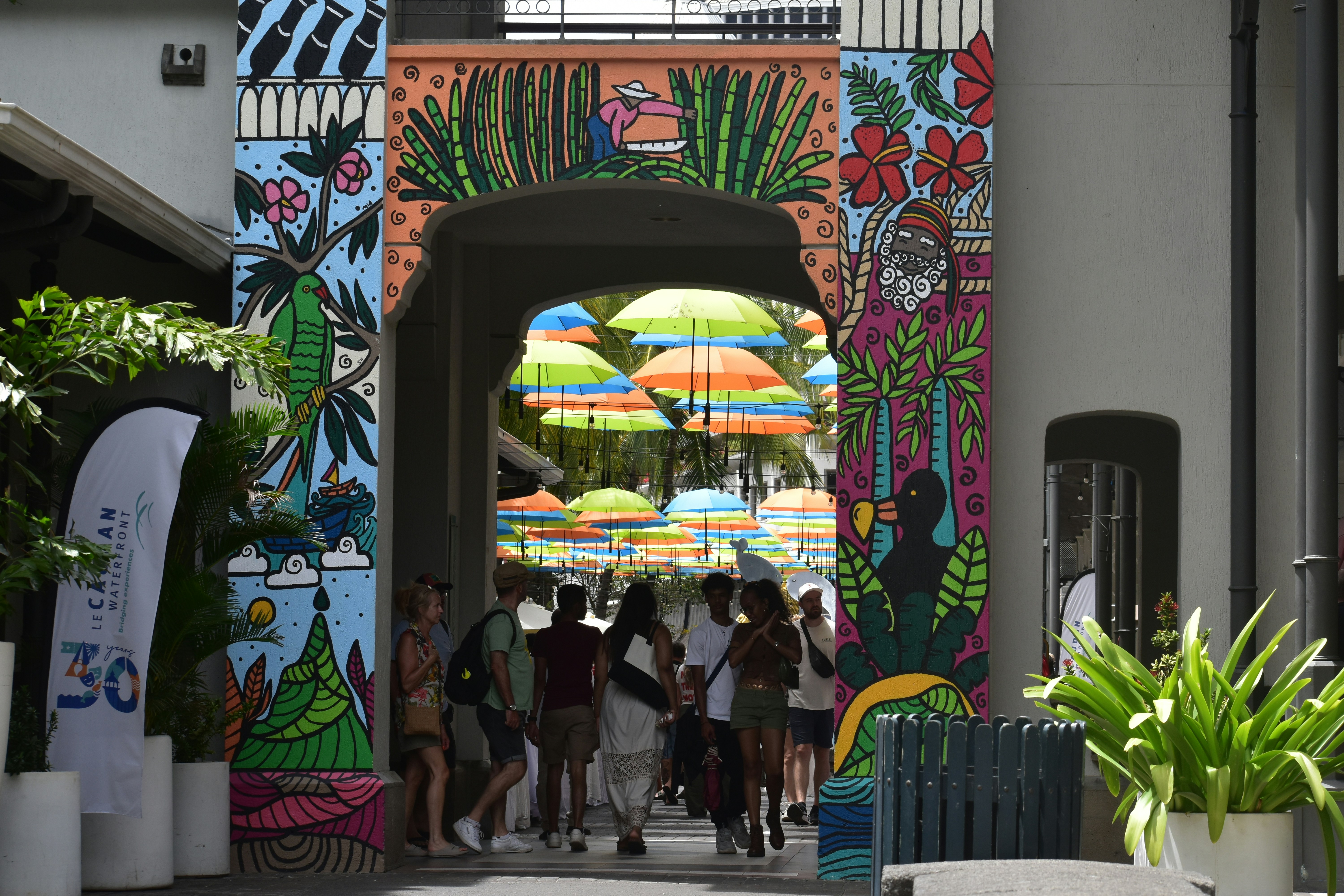 Gente caminando por un arco decorado con murales coloridos bajo paraguas colgantes