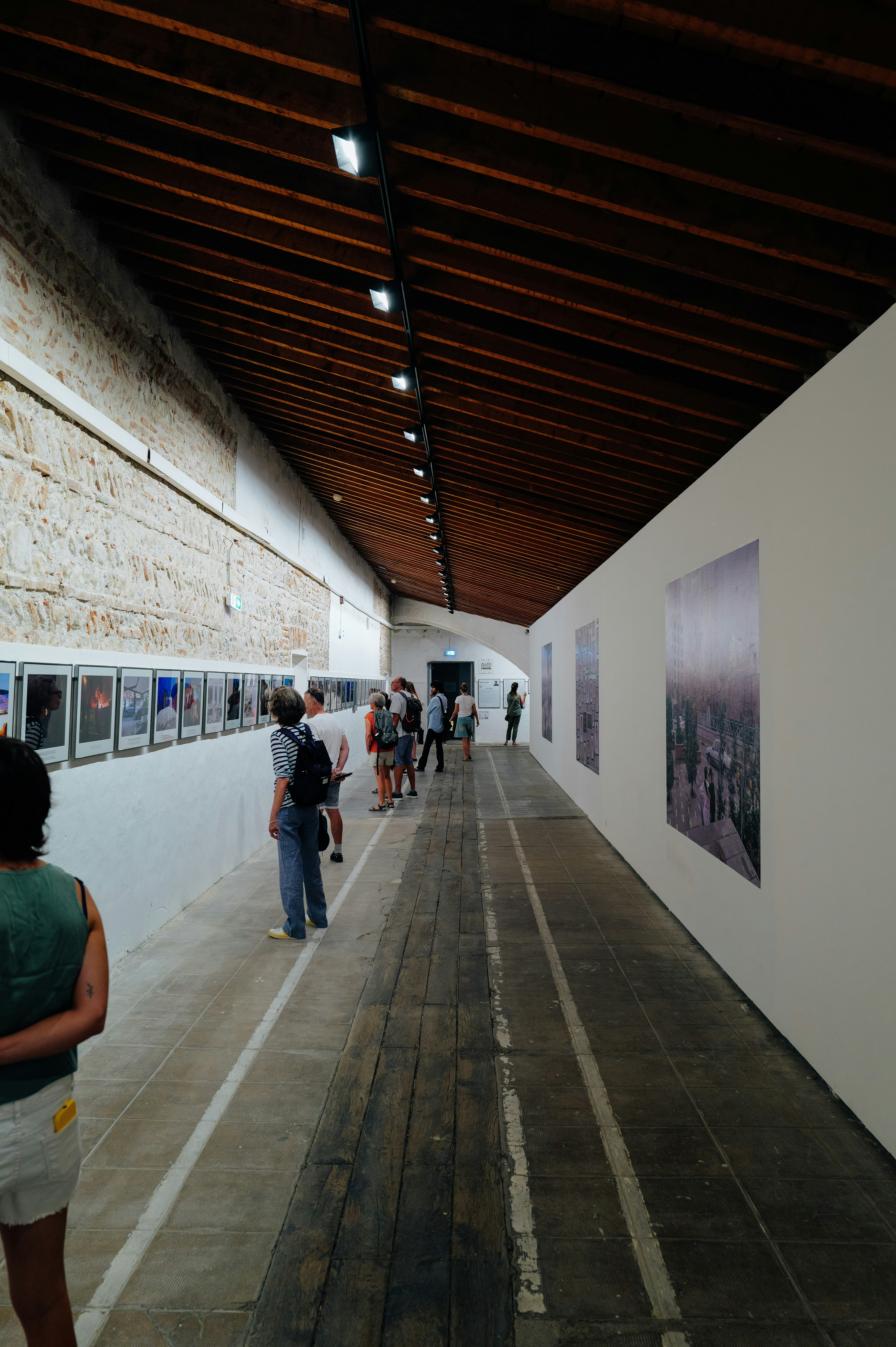 Pessoas assistindo a fotografias emolduradas ao longo de uma longa galeria com teto de madeira