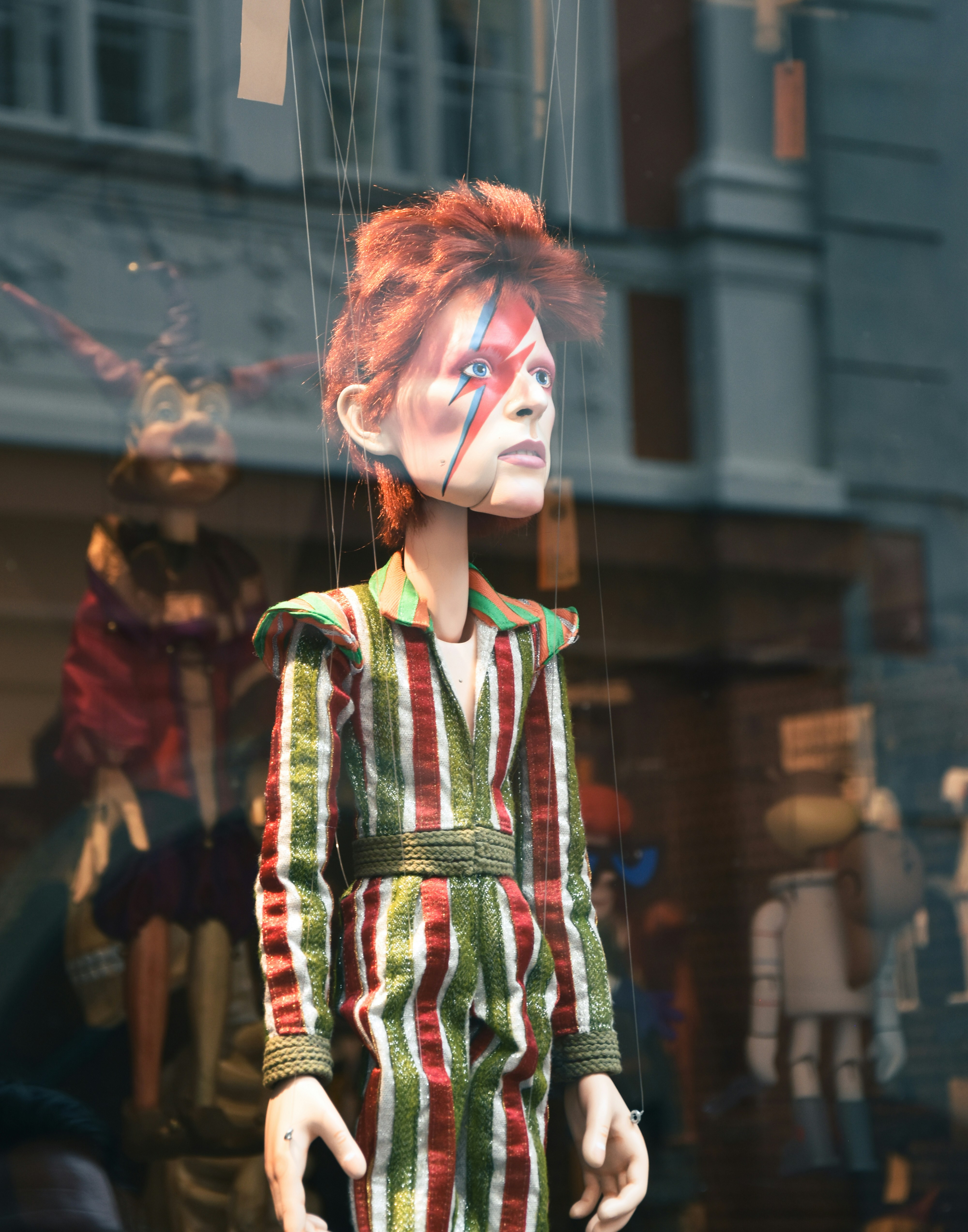 A marionette of David Bowie in a striped jumpsuit behind a glass window