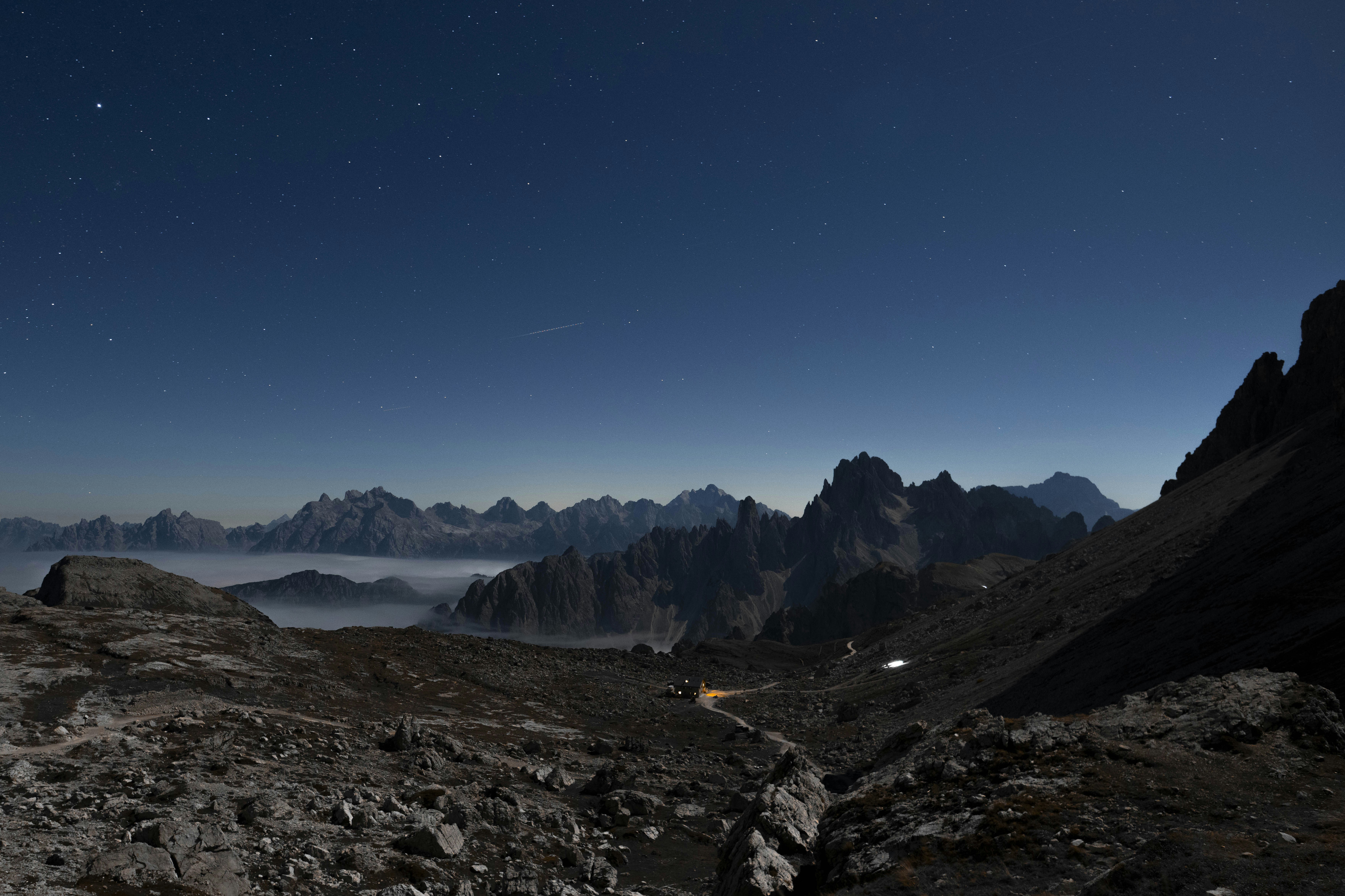 A rocky mountain landscape under a starry night sky with a glowing cabin below
