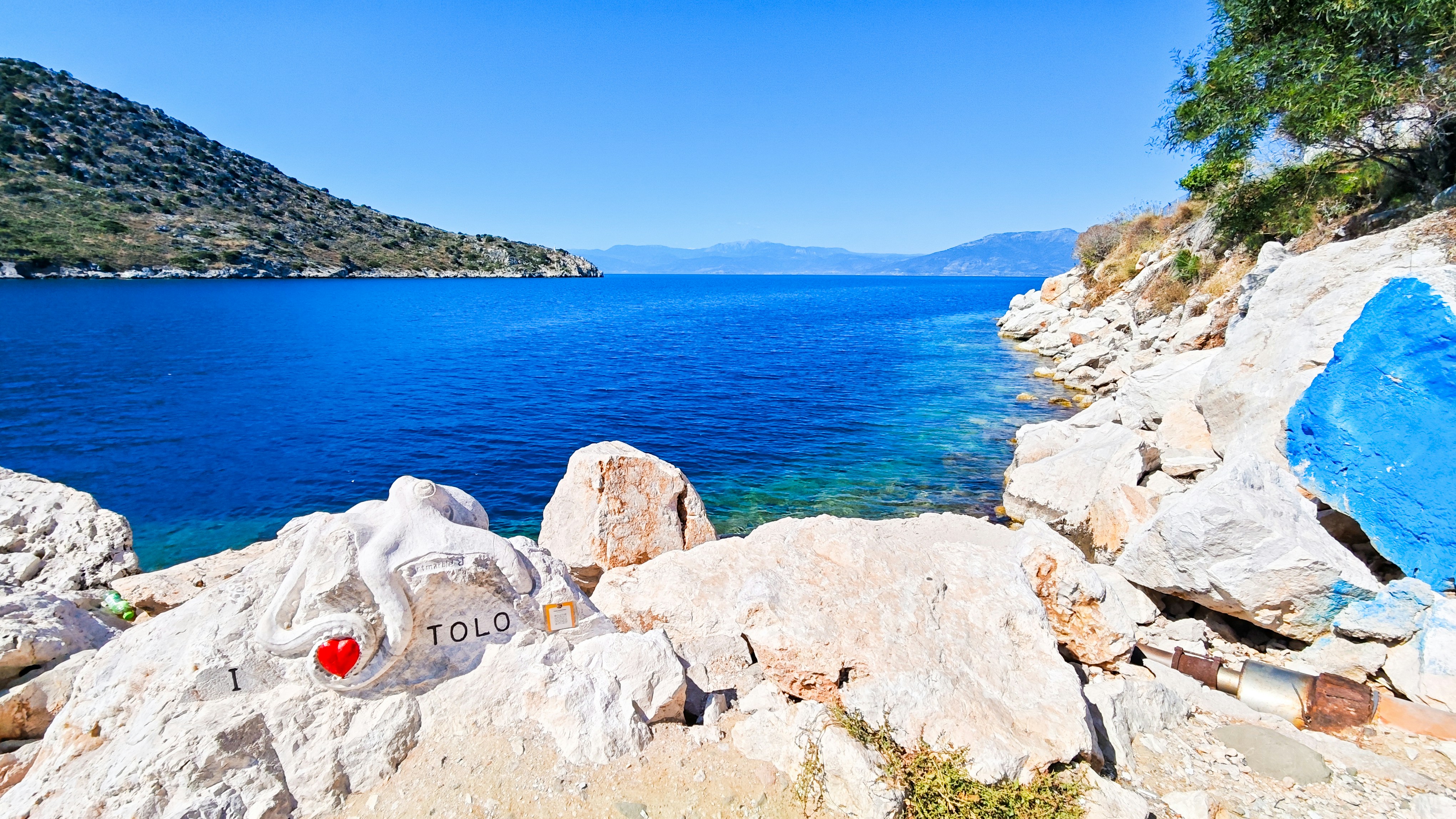 A white octopus sculpture on a rocky shore overlooking the blue sea in Tolo