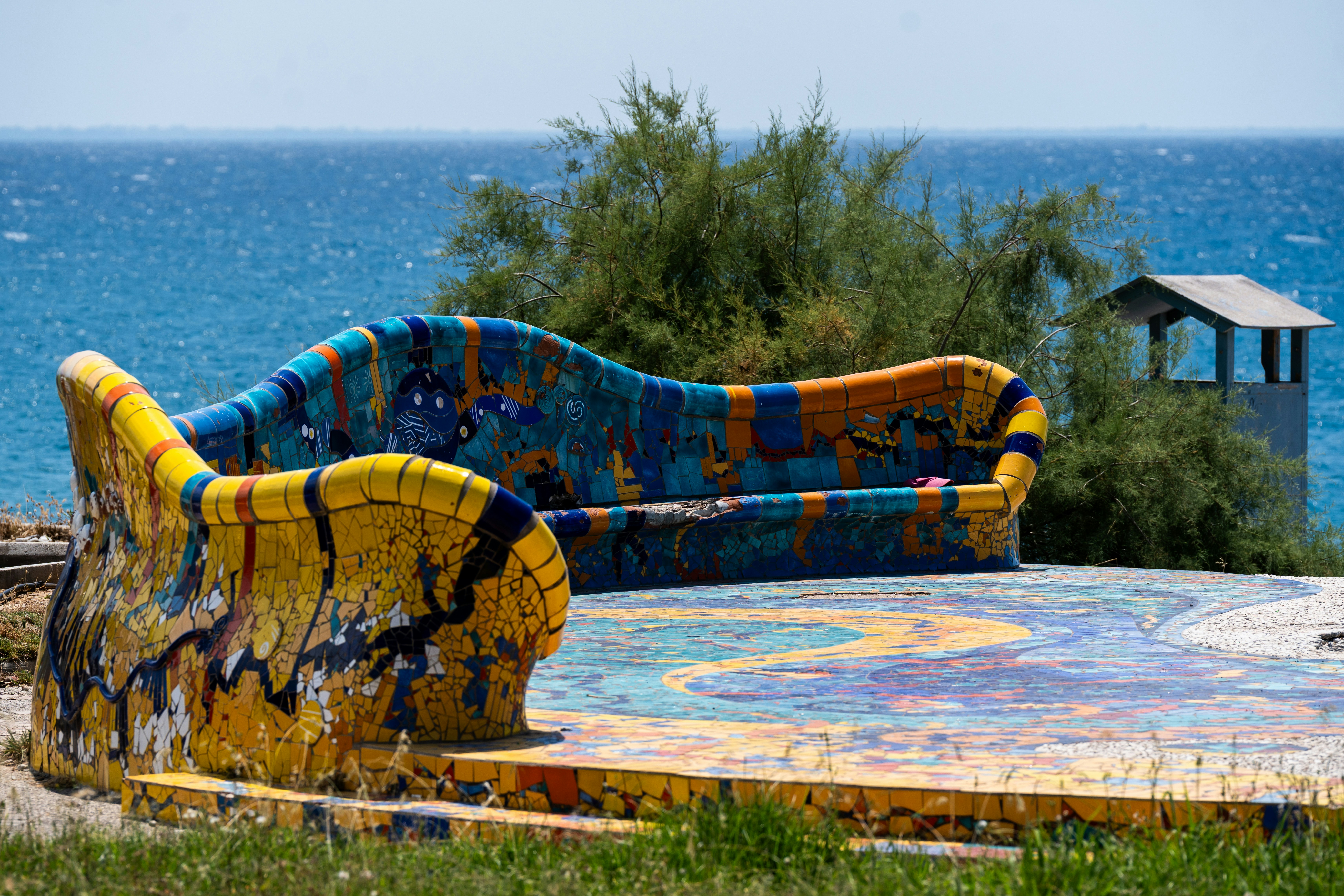 A curved mosaic bench with yellow and blue tiles overlooking the blue sea