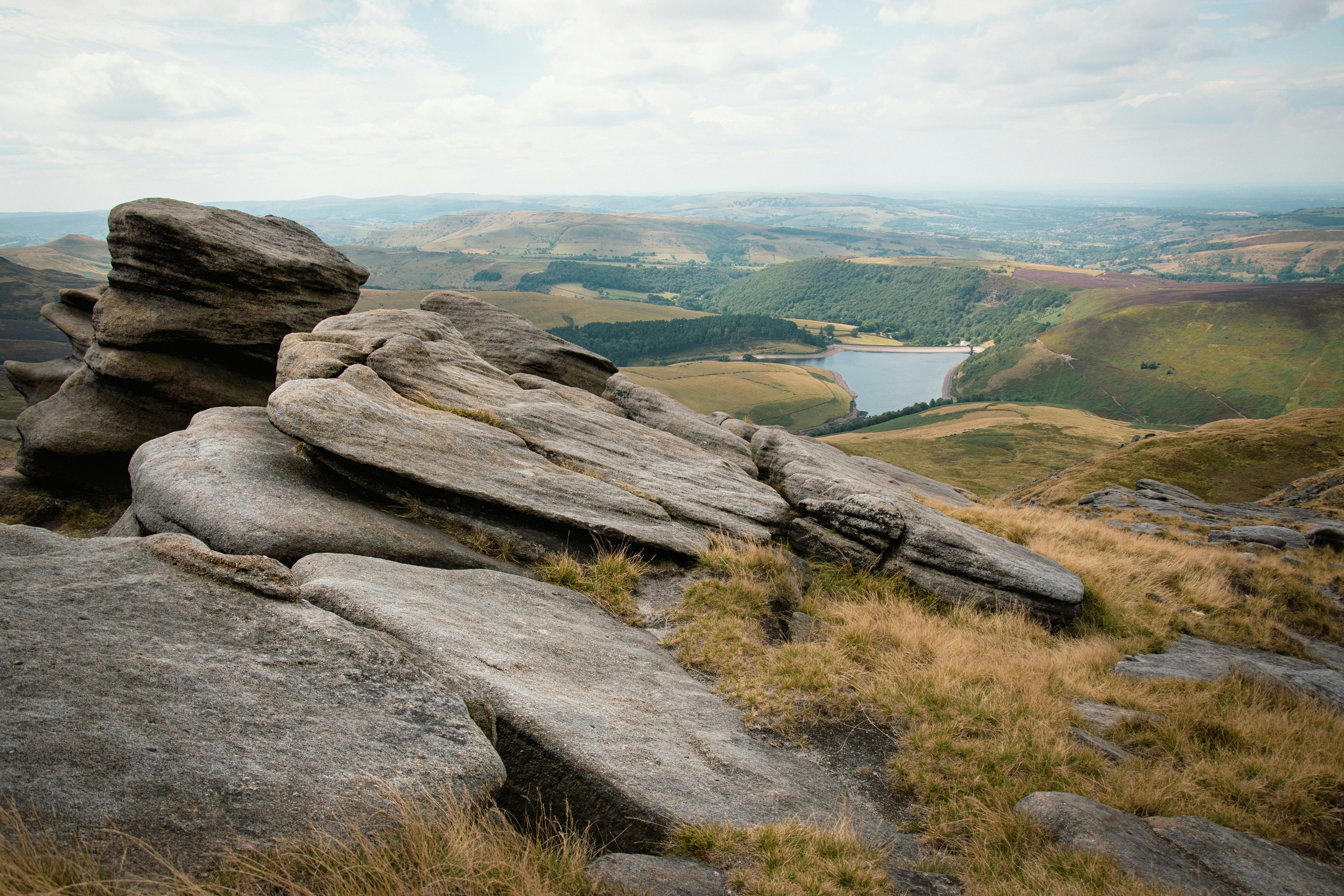 An elevated view looking down from the weathered gritstone outcrops of Kinder Scout in the Peak District. Layered rock formations in the foreground frame a sweeping vista of rolling green hills, valleys, and the Kinder Reservoir shimmering under a soft, overcast sky.