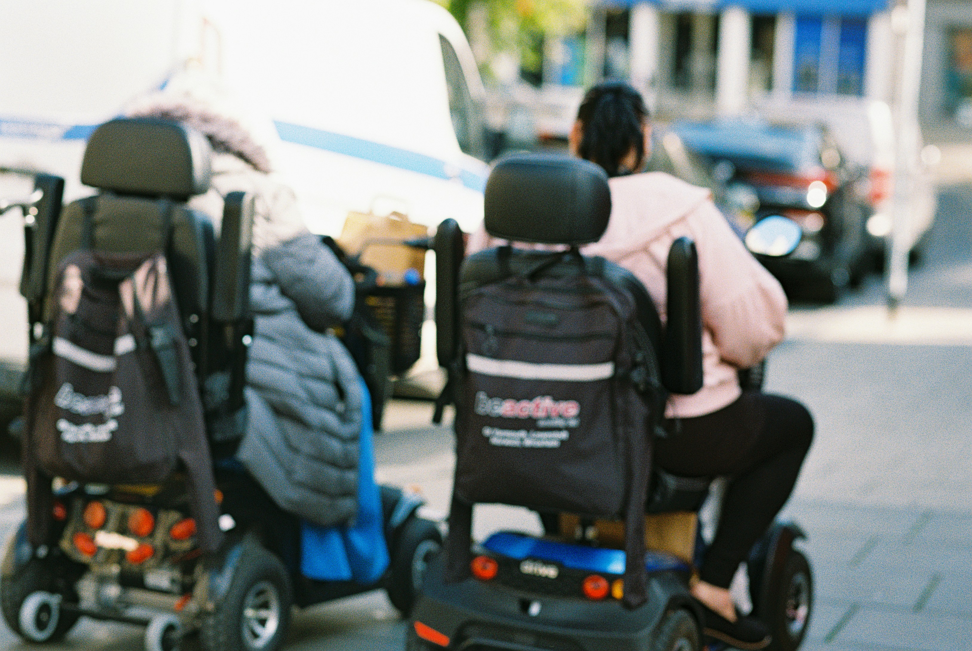 Two people on mobility scooters on a city sidewalk with a white van