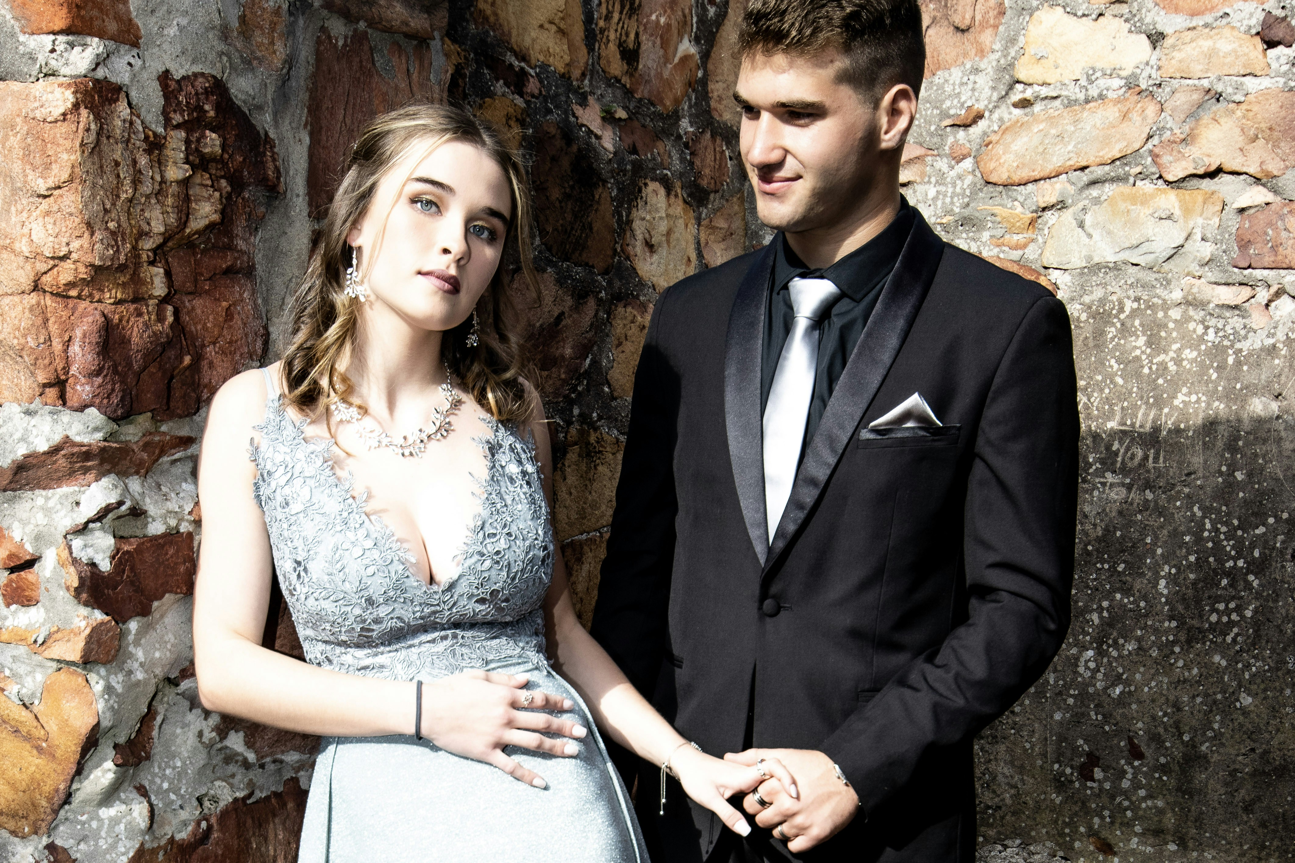 A man and a woman in formal attire holding hands against a stone wall
