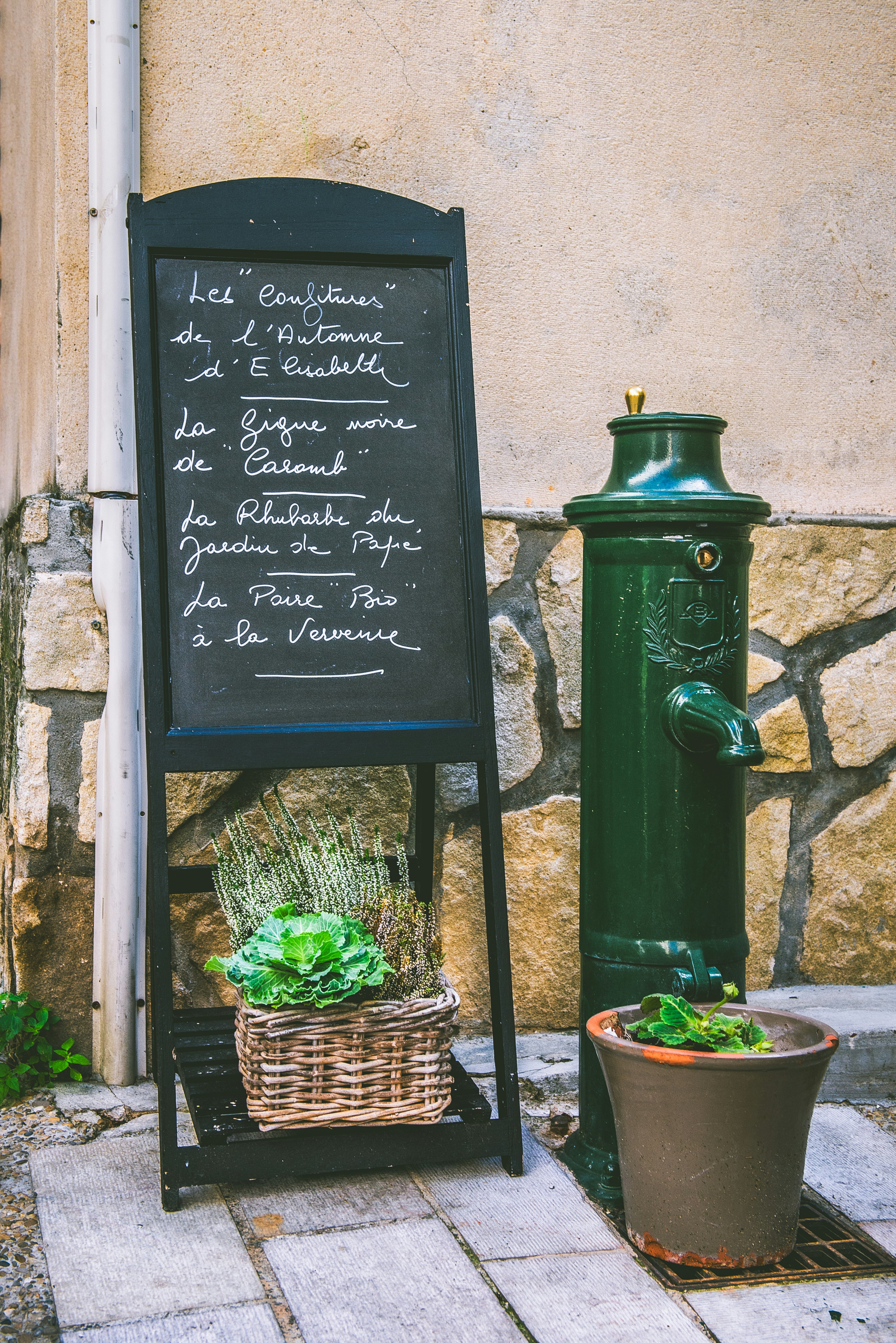 A chalkboard menu beside a green water pump and potted plants on a sidewalk