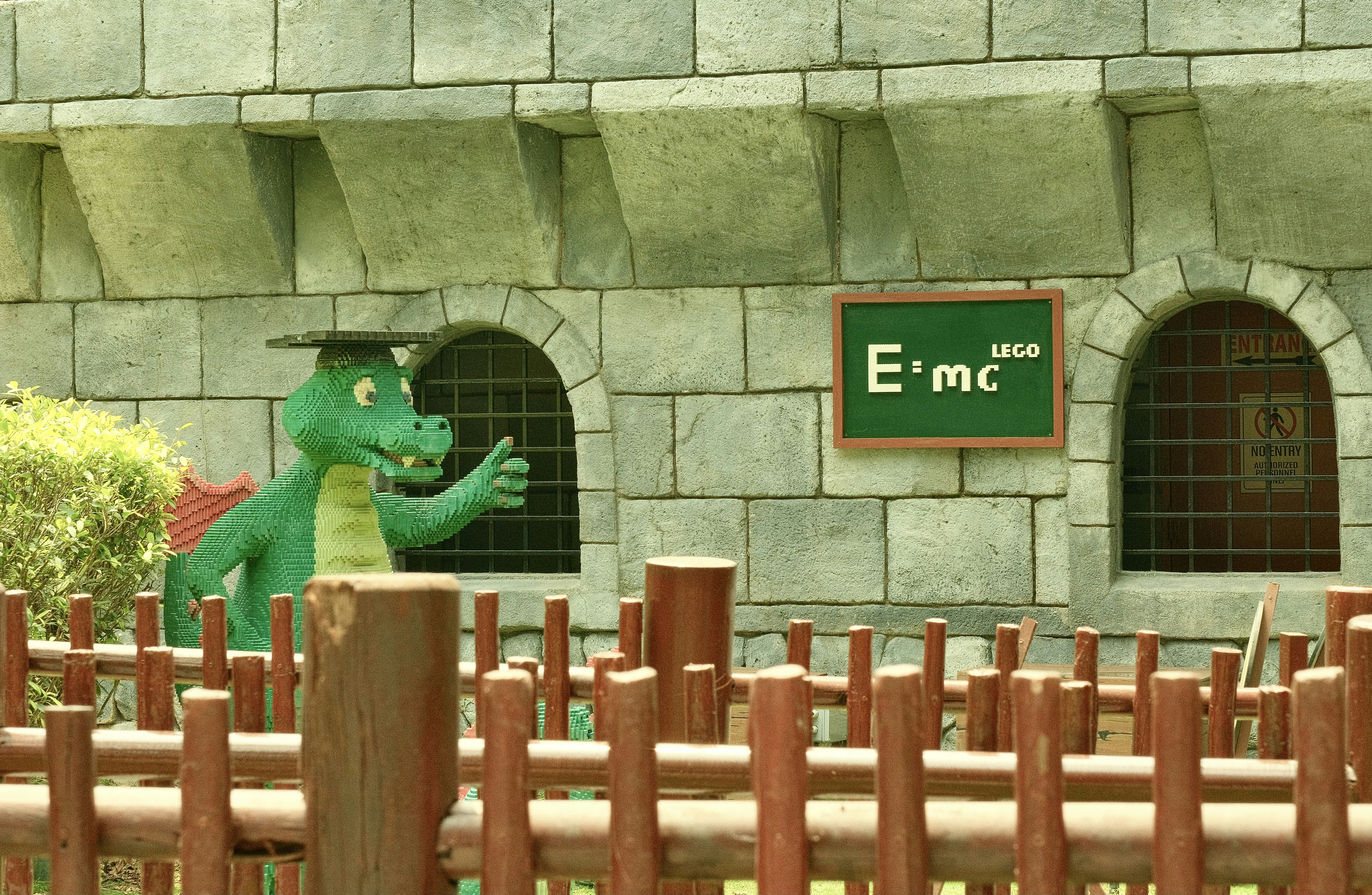 A green dragon made of plastic bricks wearing a mortarboard near a stone wall