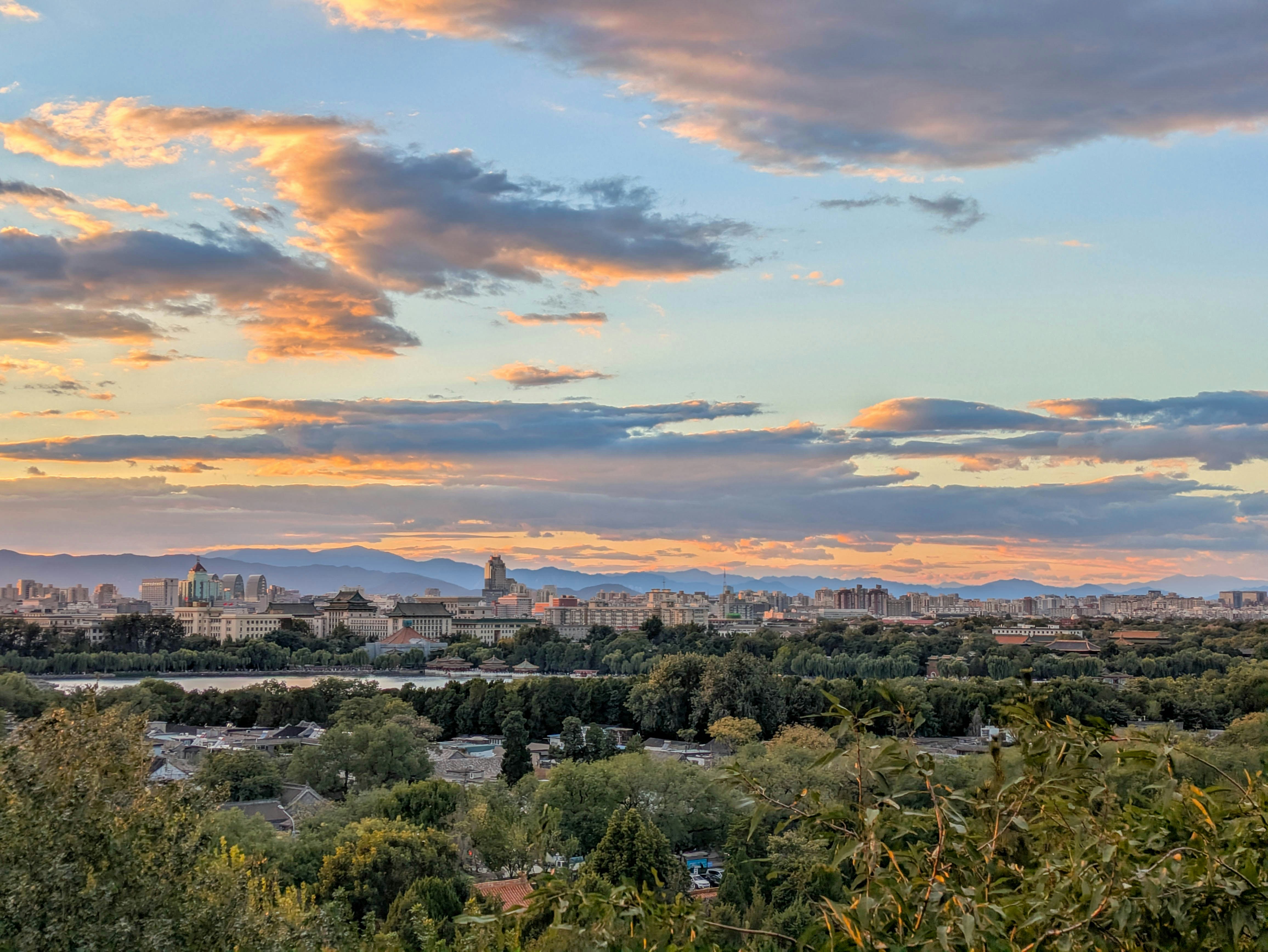 A cityscape of Beijing under a warm orange sky with clouds above trees
