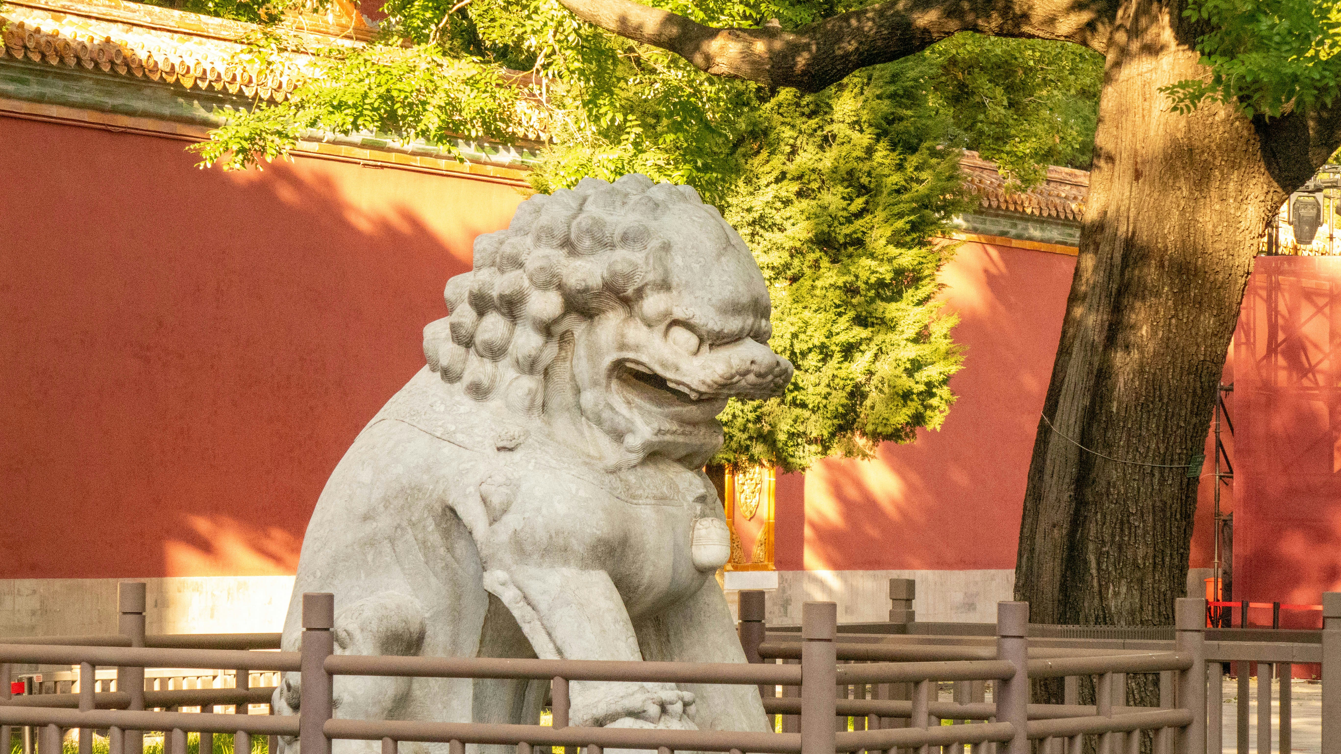 A stone guardian lion statue in front of a red wall and tree