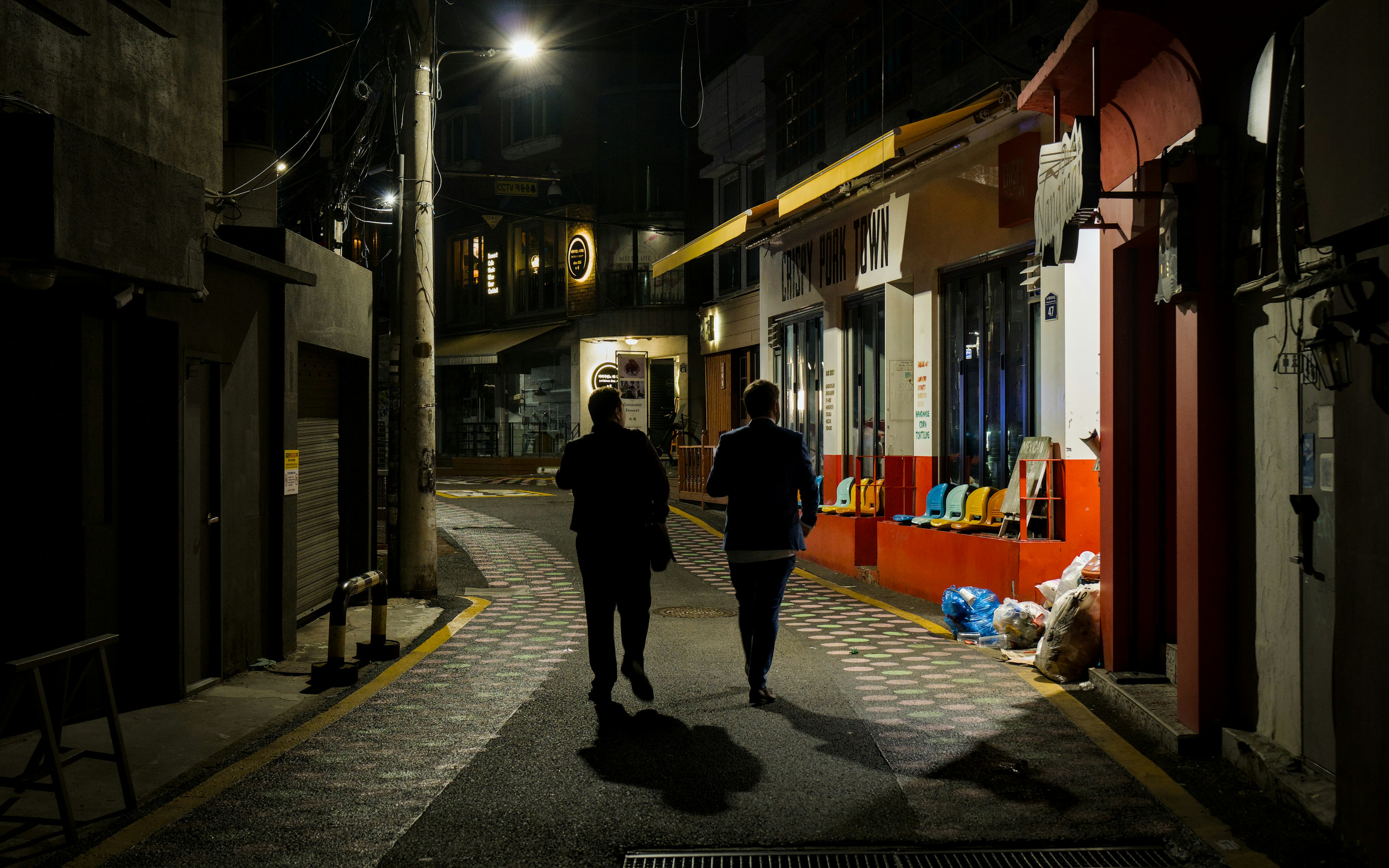 Two people walking down a narrow city street at night past a restaurant