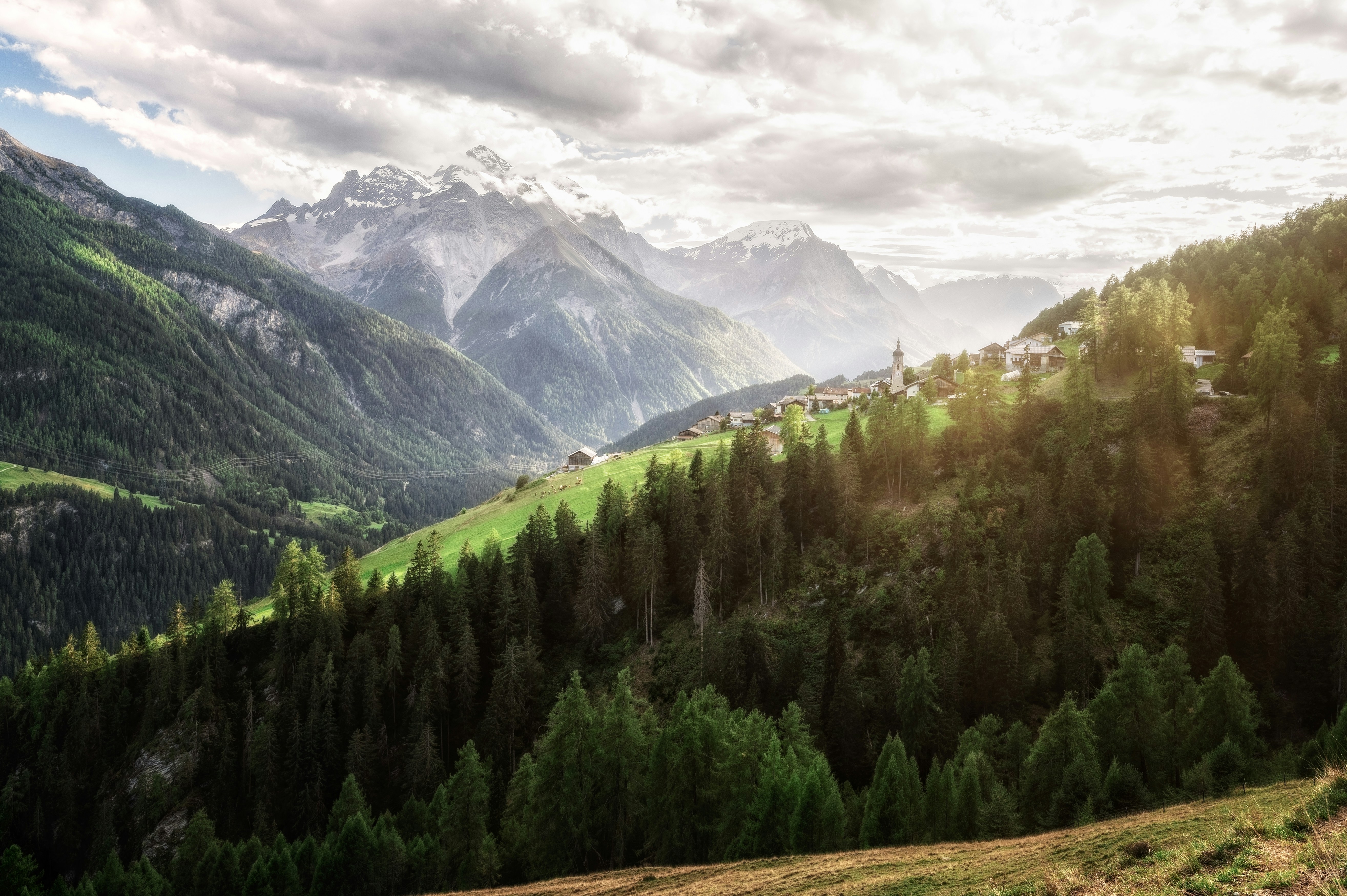 A small village on a green hillside with pine forests and distant snow-capped mountains
