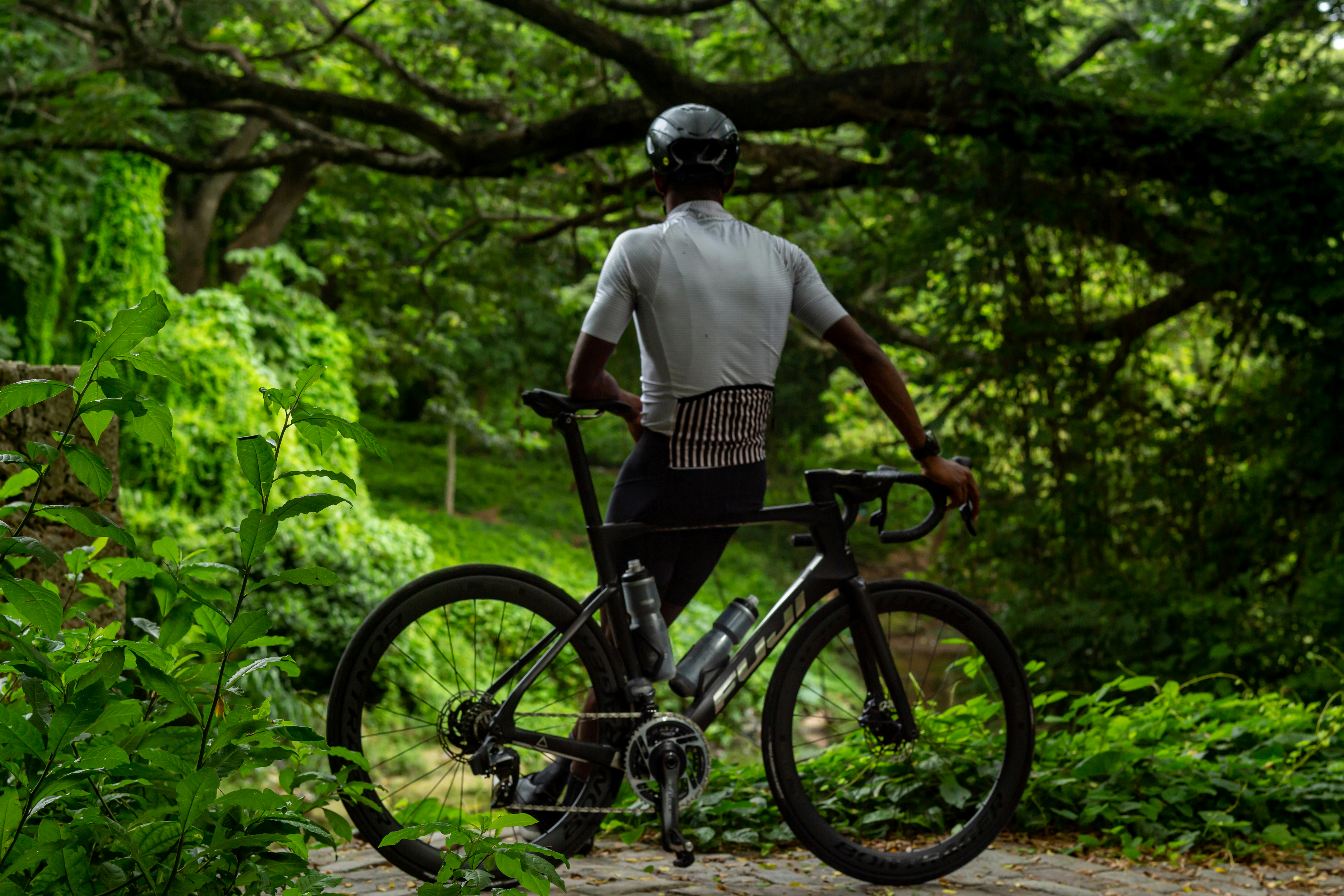 A cyclist in a helmet and jersey standing with a road bike in a forest
