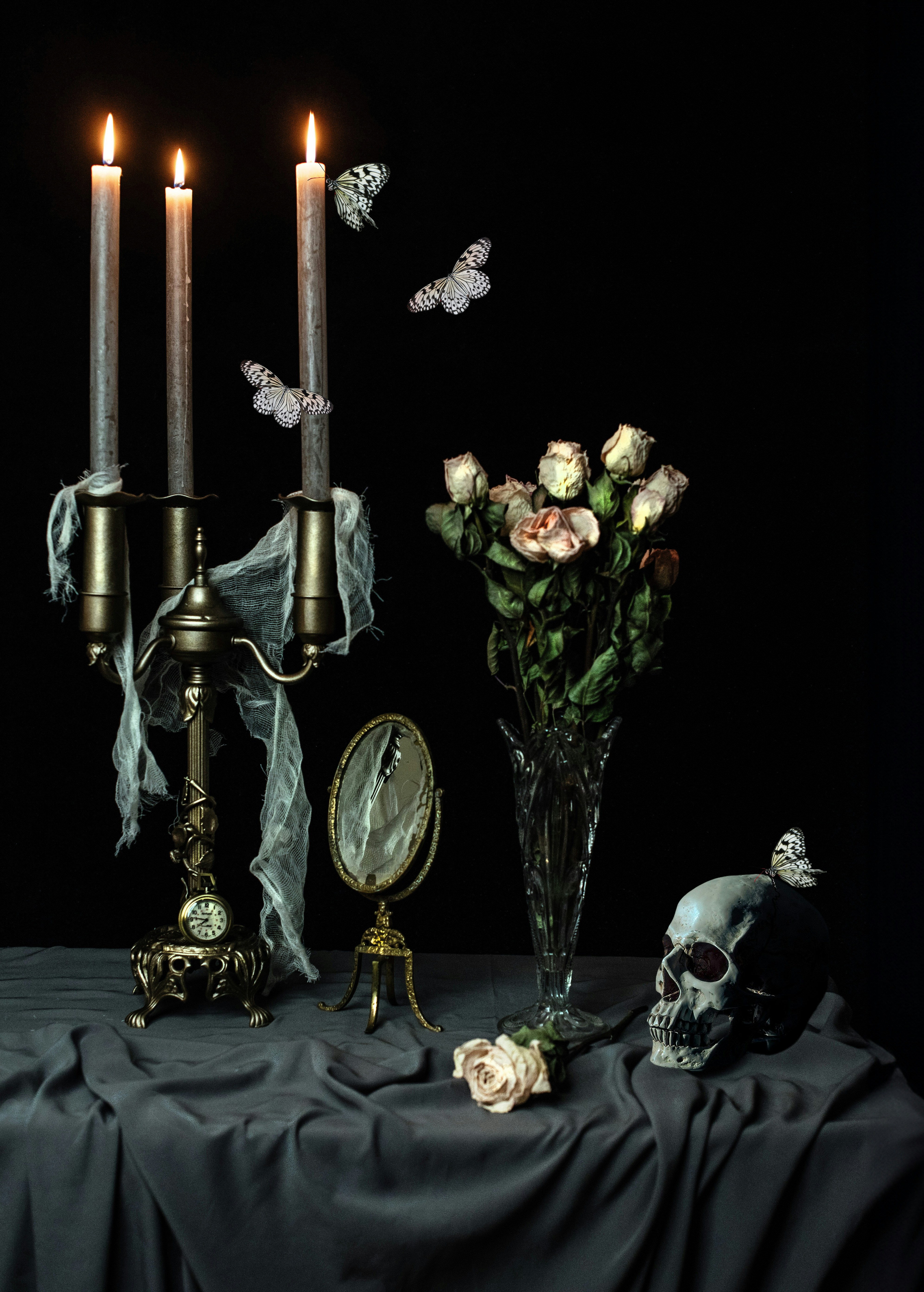 A candelabra with three lit candles, dried roses, a skull, and butterflies on a table